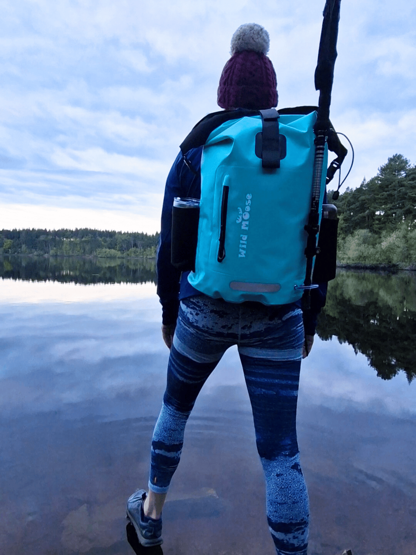lady in blue legging wearing a mint green waterproof backpack. She is standing on the edge of a lake and has her back to the camera