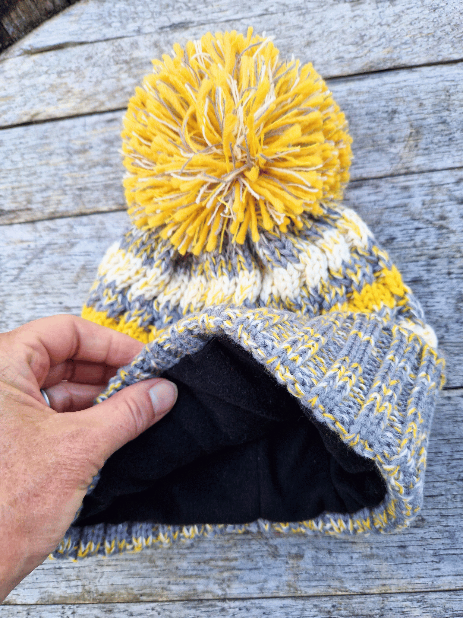 Knitted beanie with yellow pom-pom held by a hand to show fleece lining. on a wooden surface