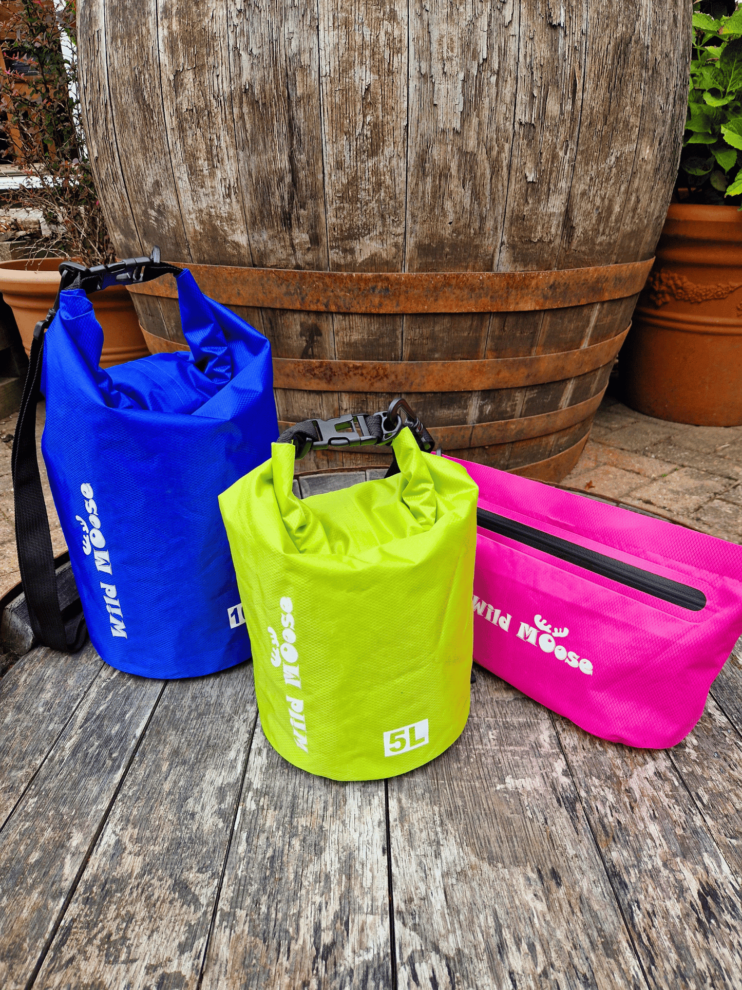 Three colorful dry bags (blue, green, pink) on a wooden surface with a wooden barrel in the background.