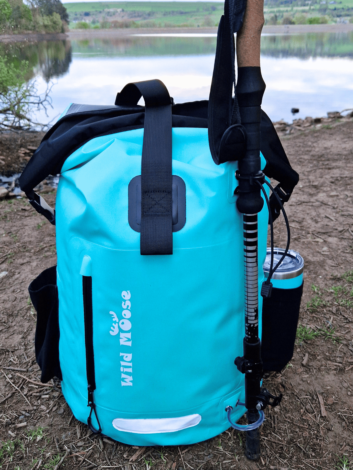 a minty green backpack on the ground in front of a reservoir. A flask in the side pocket and a single walking pole on the front