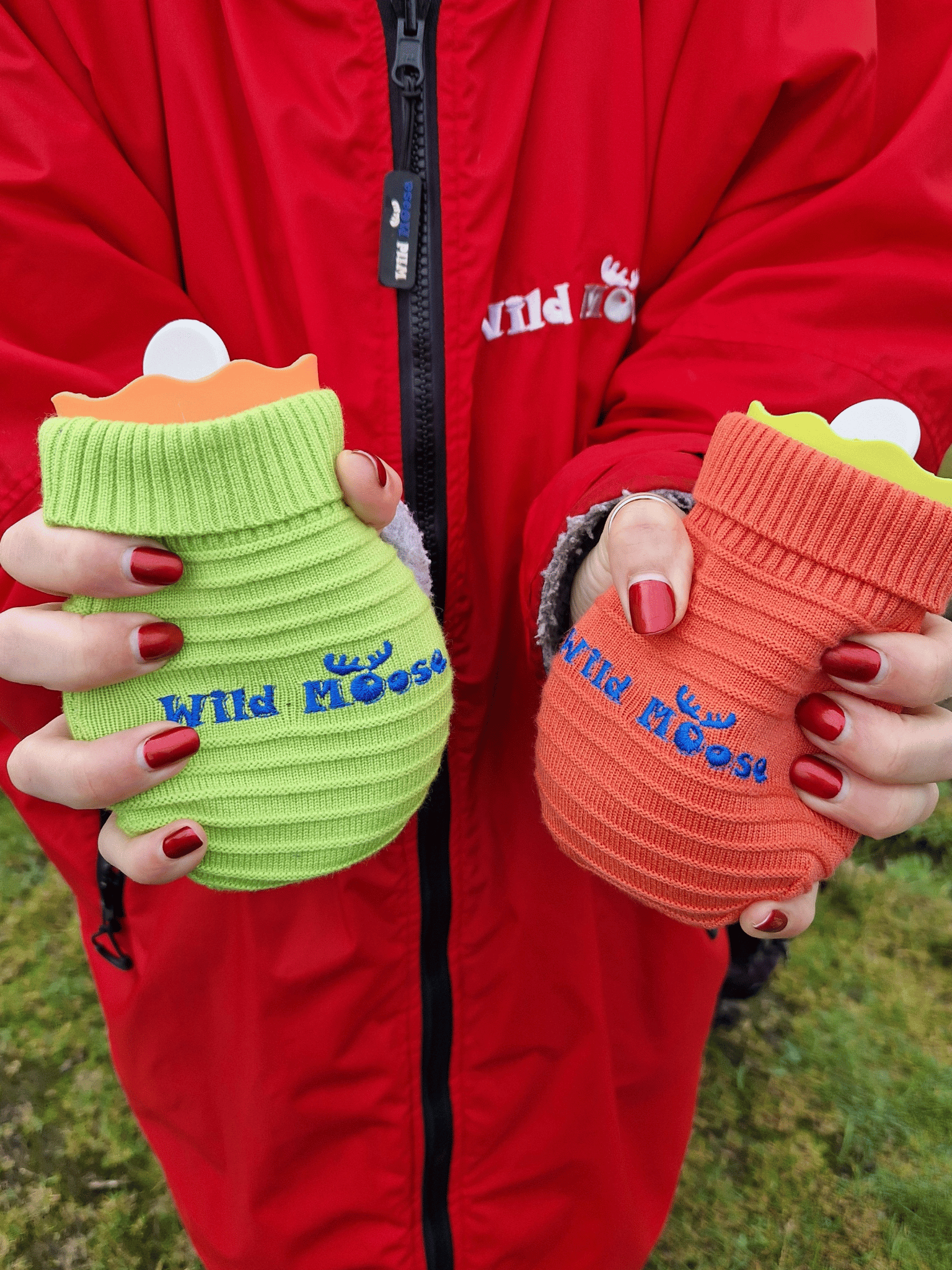brightly coloured mini hot water bottles in a knitted cover in orange and lime green - held by a lady wearing a red changing robe and with red nail polish on her fingernails