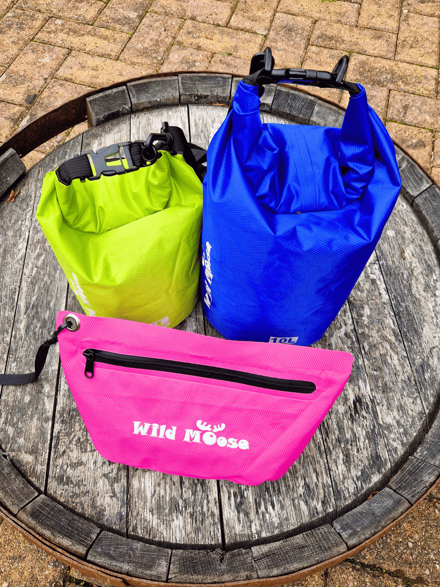 Three waterproof bags in blue, green, and pink on a wooden surface with a brick background.