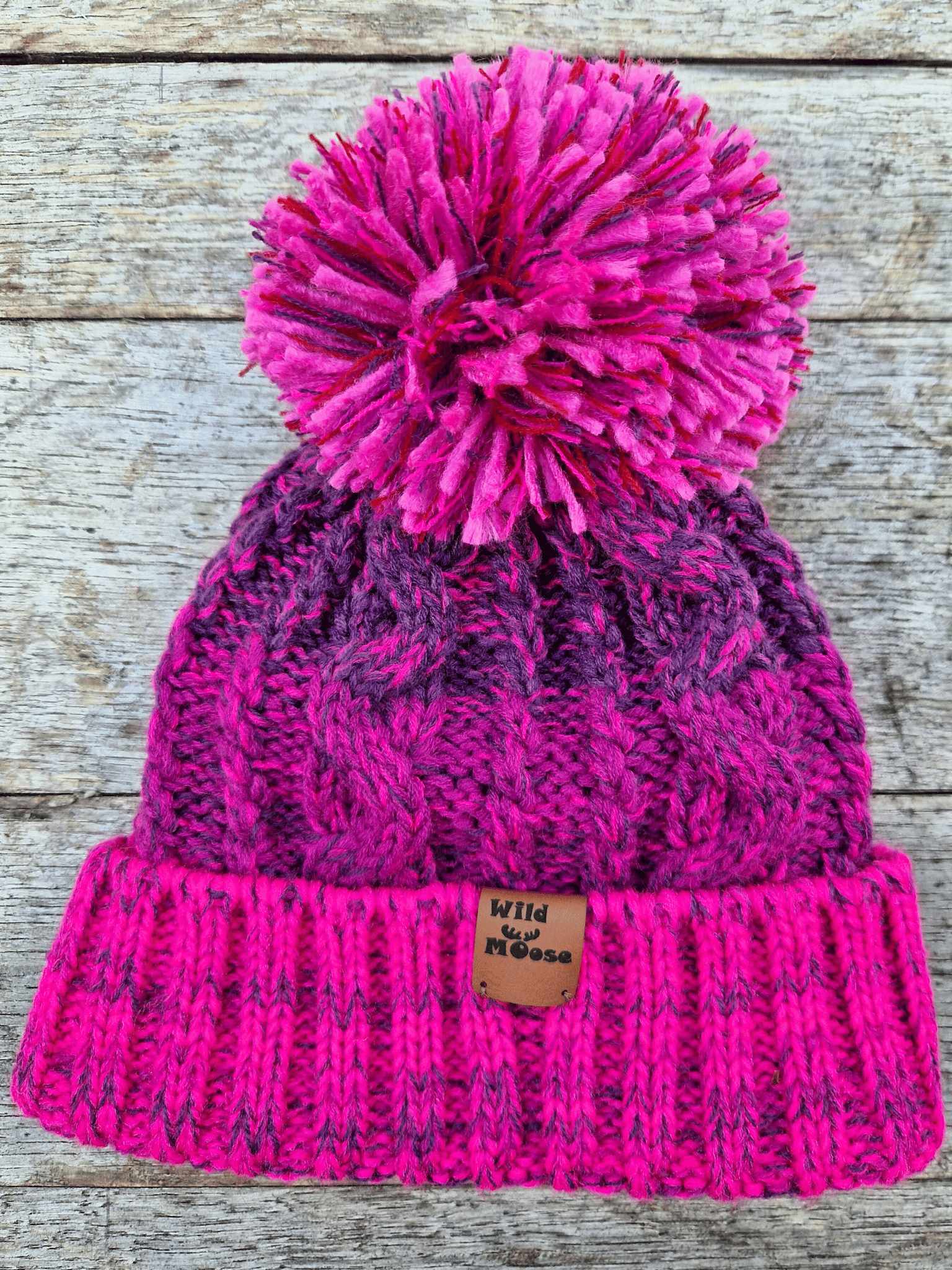 Purple and pink knit beanie with a pom-pom on a wooden surface
