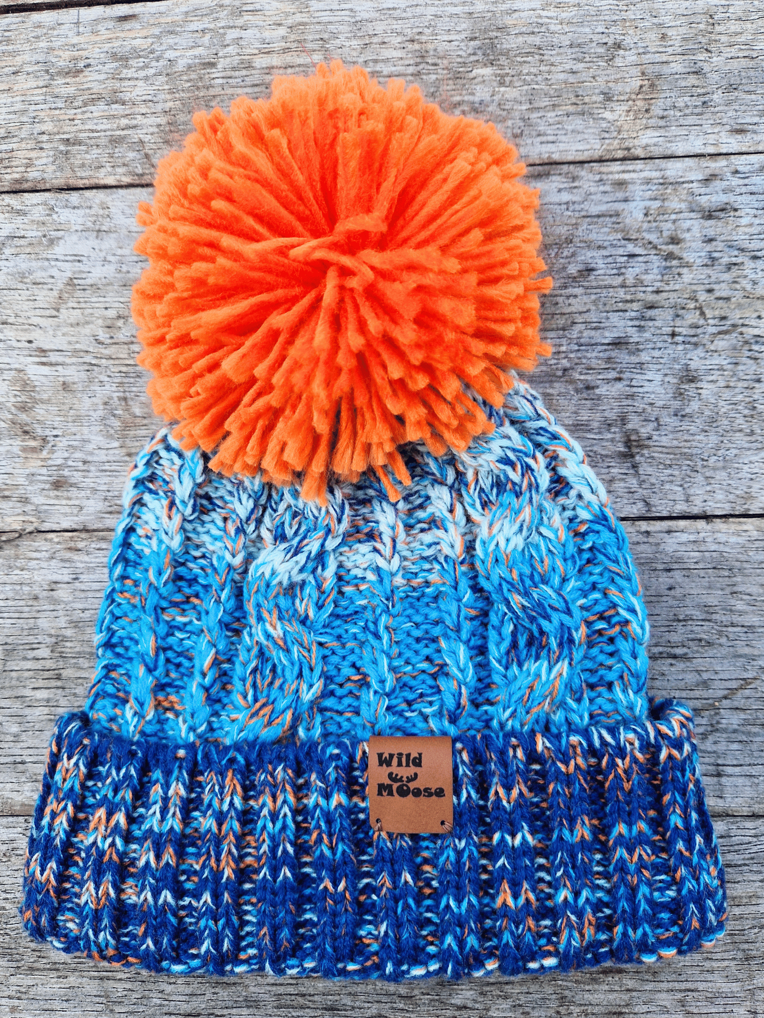 Blue knit beanie with orange pom-pom on a wooden surface