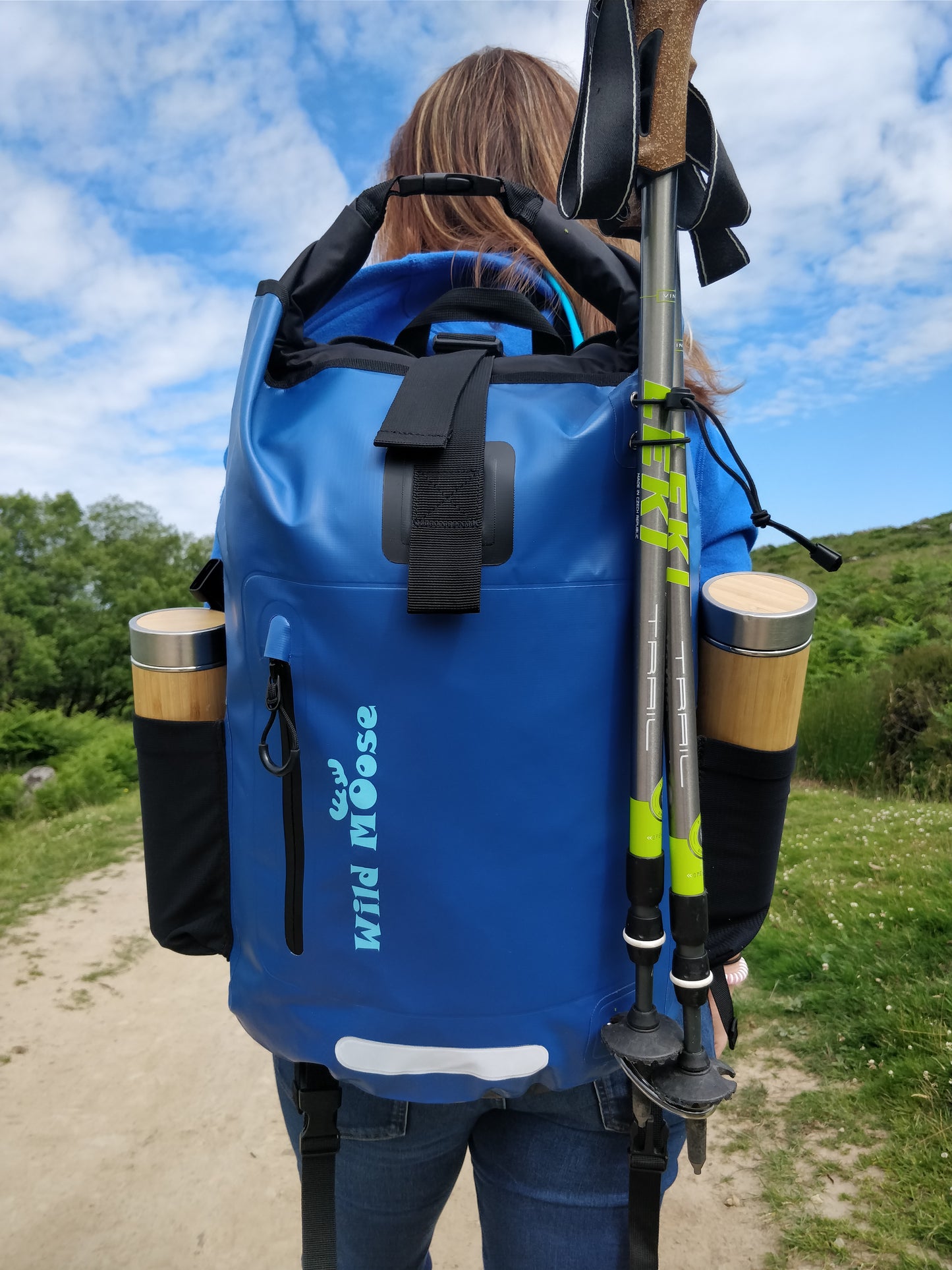 a lady walking up a path with a bright blue waterproof backpack on her back. She has 2 flasks in the side pockets and 2 walking poles attached