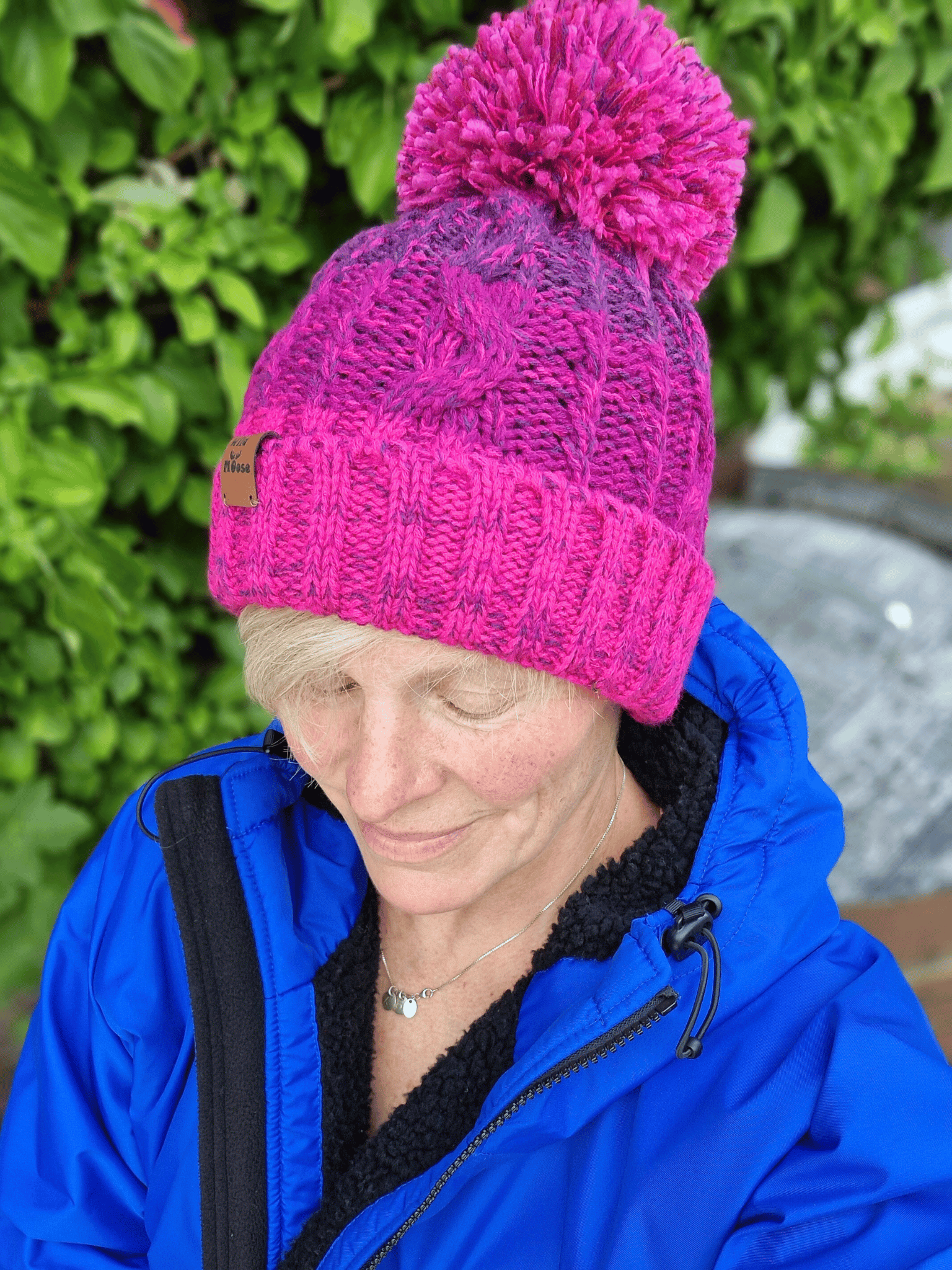 Person wearing a bright pink knit beanie with a large pom-pom, standing outdoors in an electric blue changing robe.