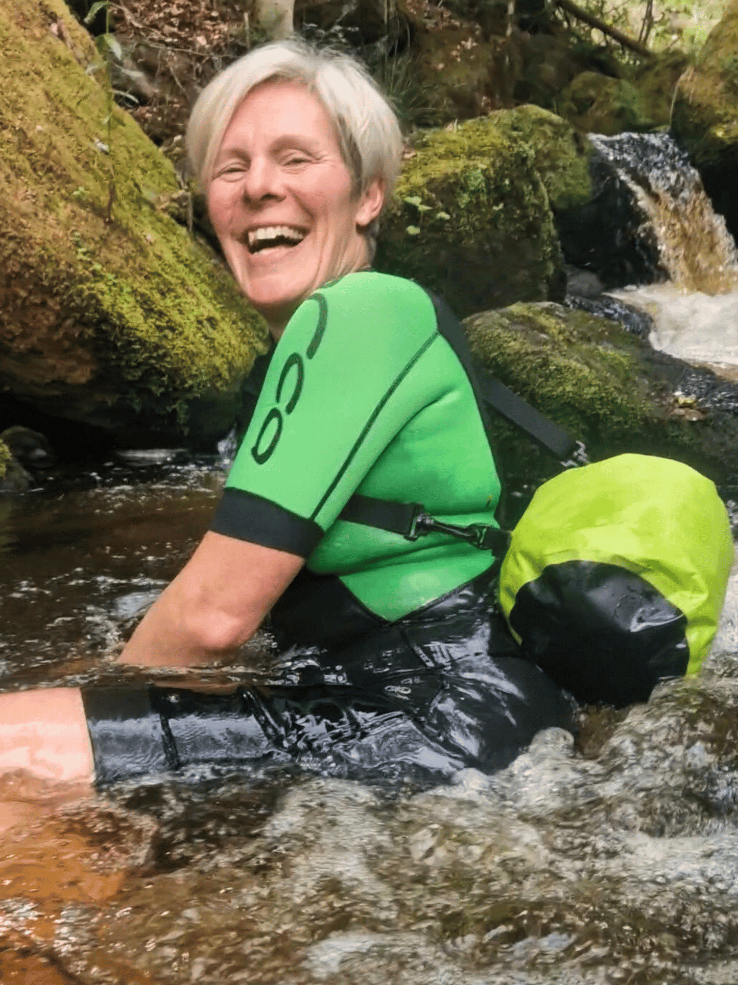 lady in water laughing, wearing a wetsuit and with 5L neon green bag in the water behind her