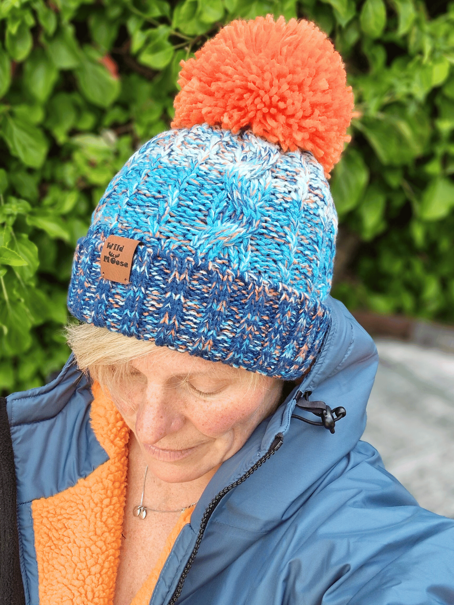Person wearing a blue and orange knit beanie with a visible brand logo, standing against a green leafy background.