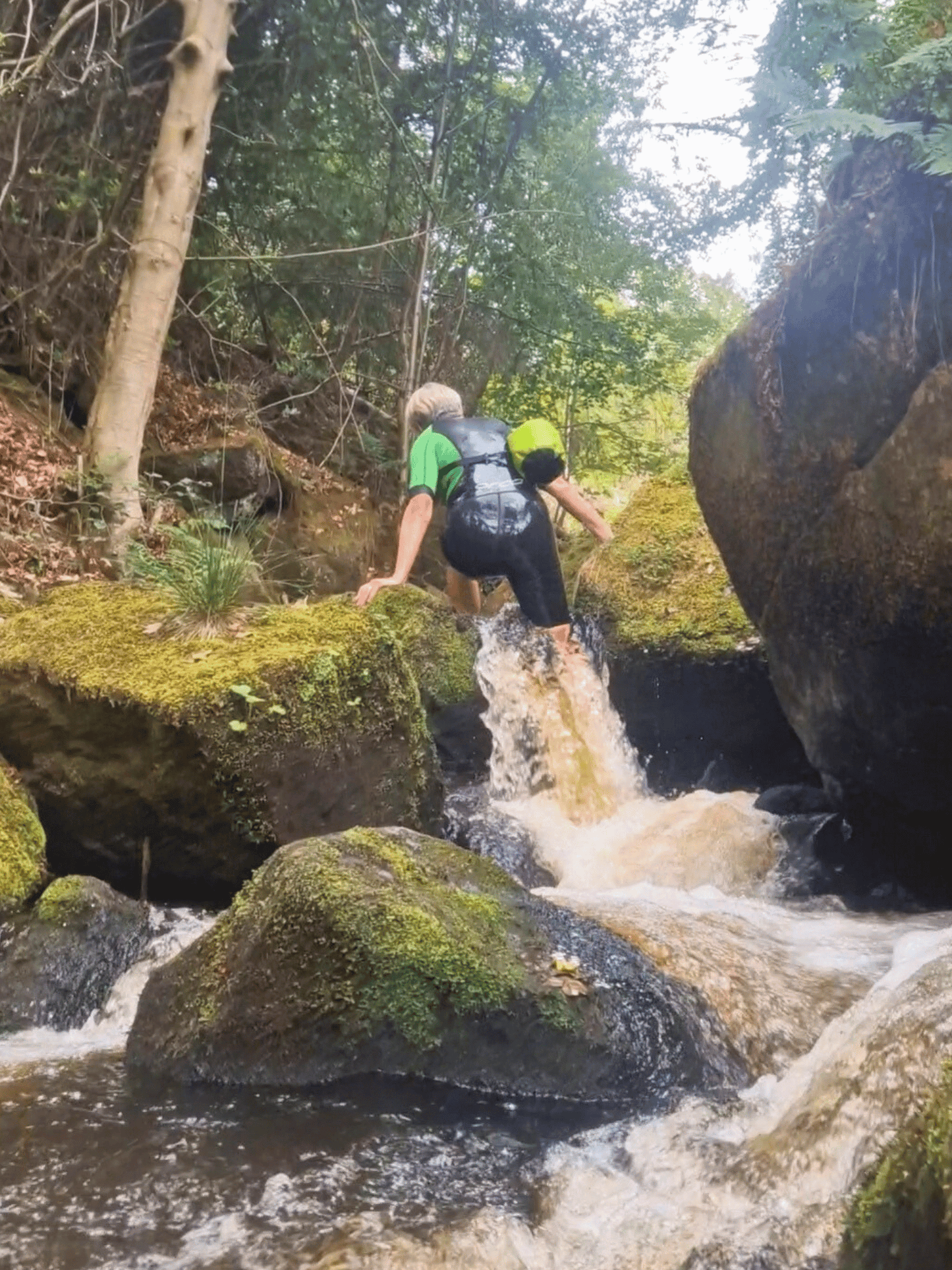 lady in wetsuit stepping out of small waterfall. She has green 5L dry bag over shoulder