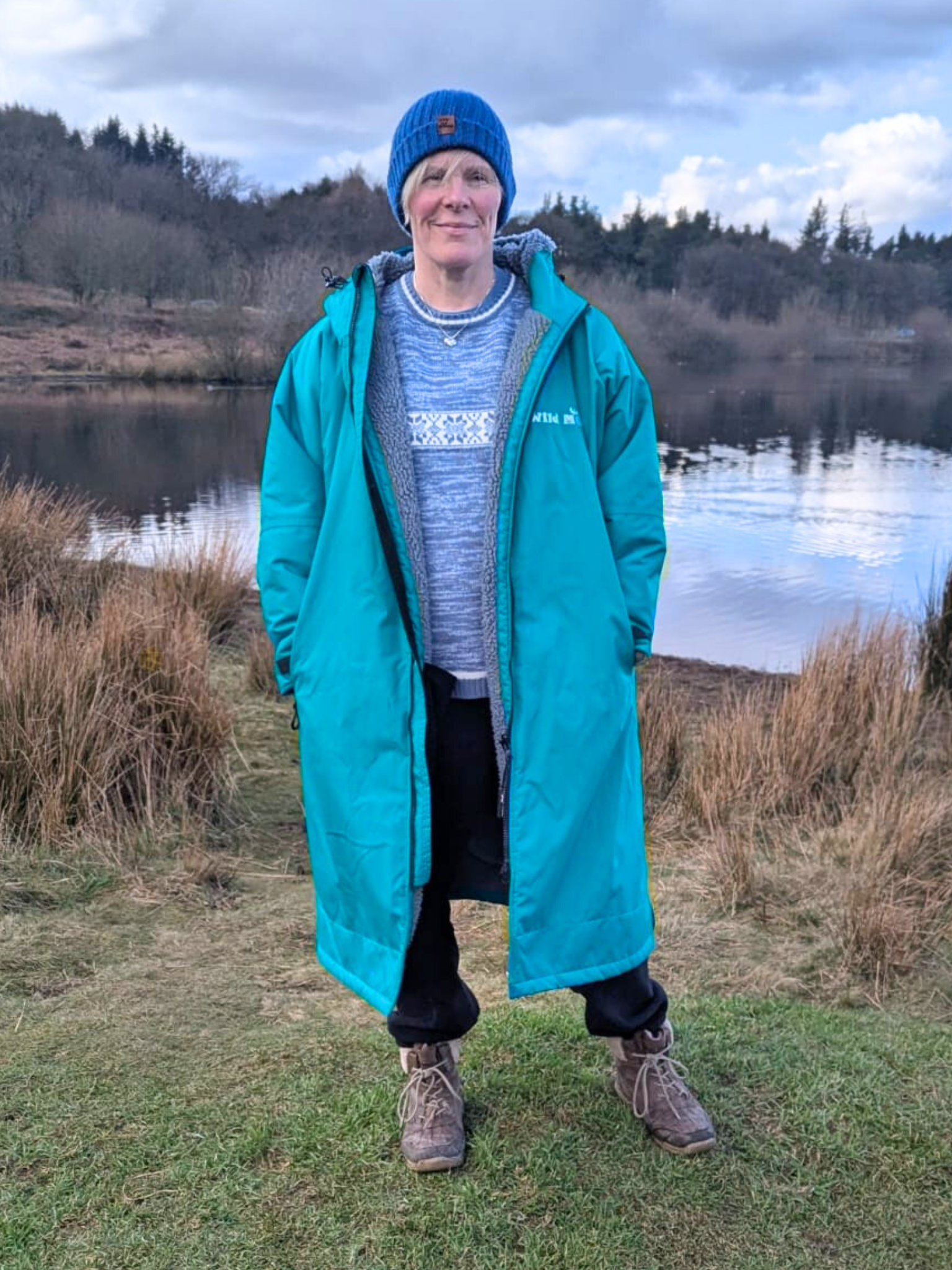 lady in teal green changing robe with hands in pockets stood by a lake. The robe has a grey fleece lining