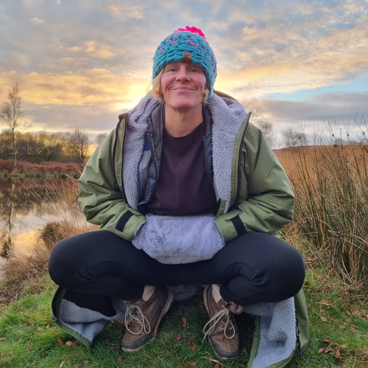 a lady is squatting by a lake in front of a sun set sky. She is wearing a grey faux fur hottum, a khaki changing robe with a grey lining and a bright colour bobble hat with a big pompom