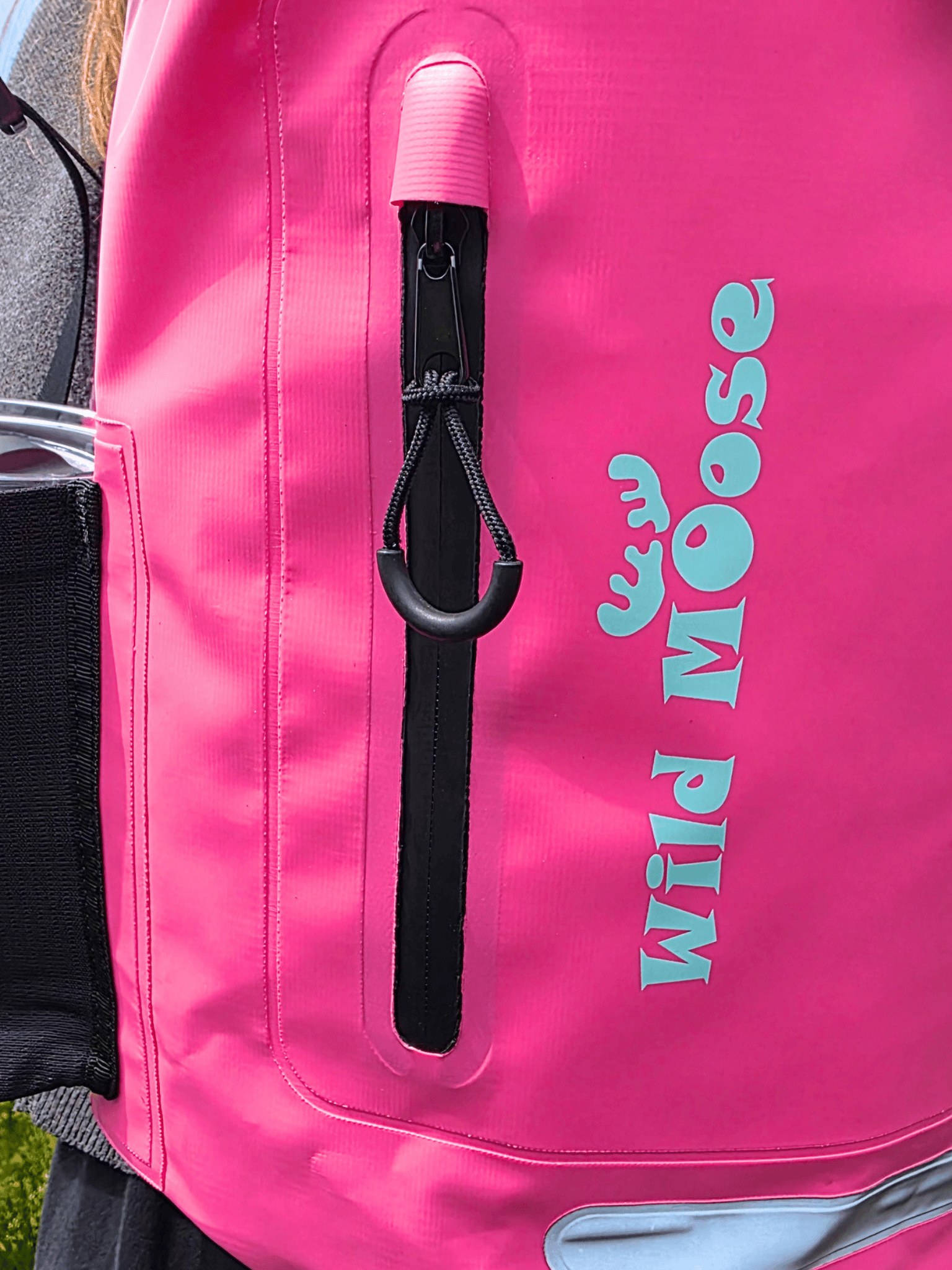 Close-up of a bright pink backpack with a vertical black zipper pocket, and turquoise 'WILD MOOSE' logo printed on the side.