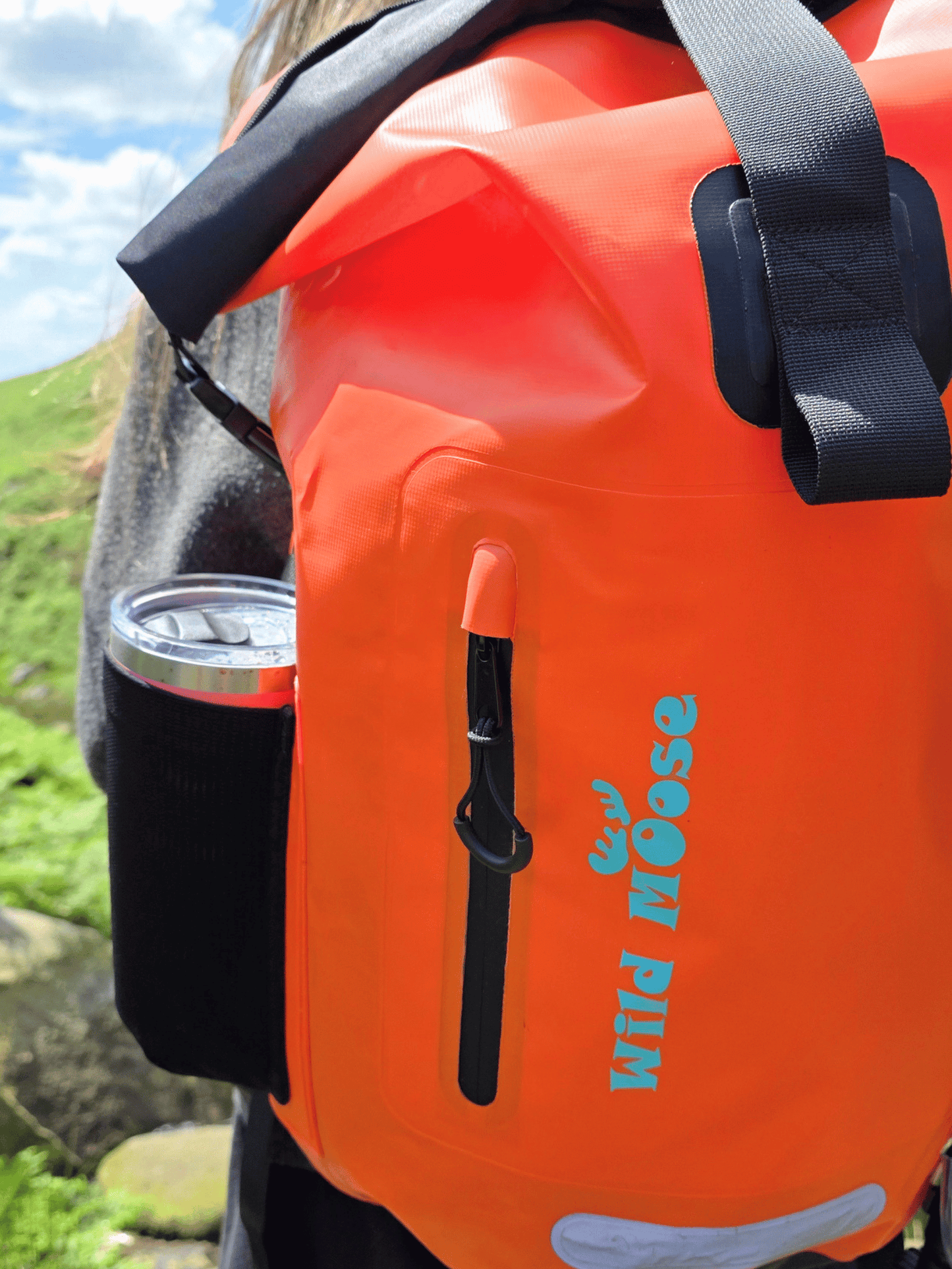Bright orange waterproof backpack with black straps and a black side cup holder, shown outdoors on a person's back. The front has a vertical zipper pocket with a black pull and the brand name 'Wild Goose' printed in teal.