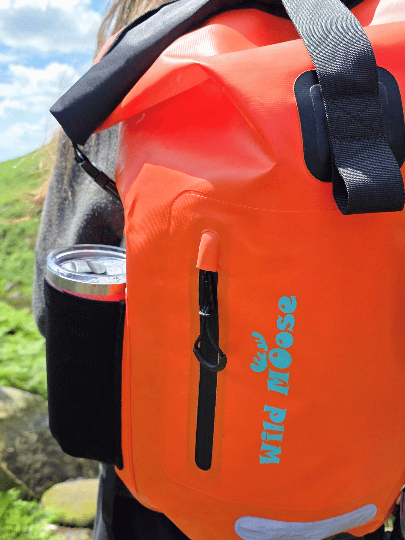 Bright orange waterproof backpack with black straps and a black side cup holder, shown outdoors on a person's back. The front has a vertical zipper pocket with a black pull and the brand name 'Wild Goose' printed in teal.