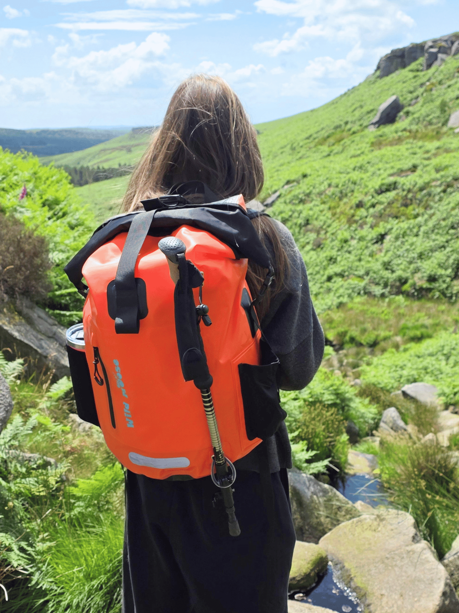 A person viewed from behind wearing a bright orange waterproof backpack with black straps and gear, standing outdoors on a grassy mountainside near a stream under a blue sky.
