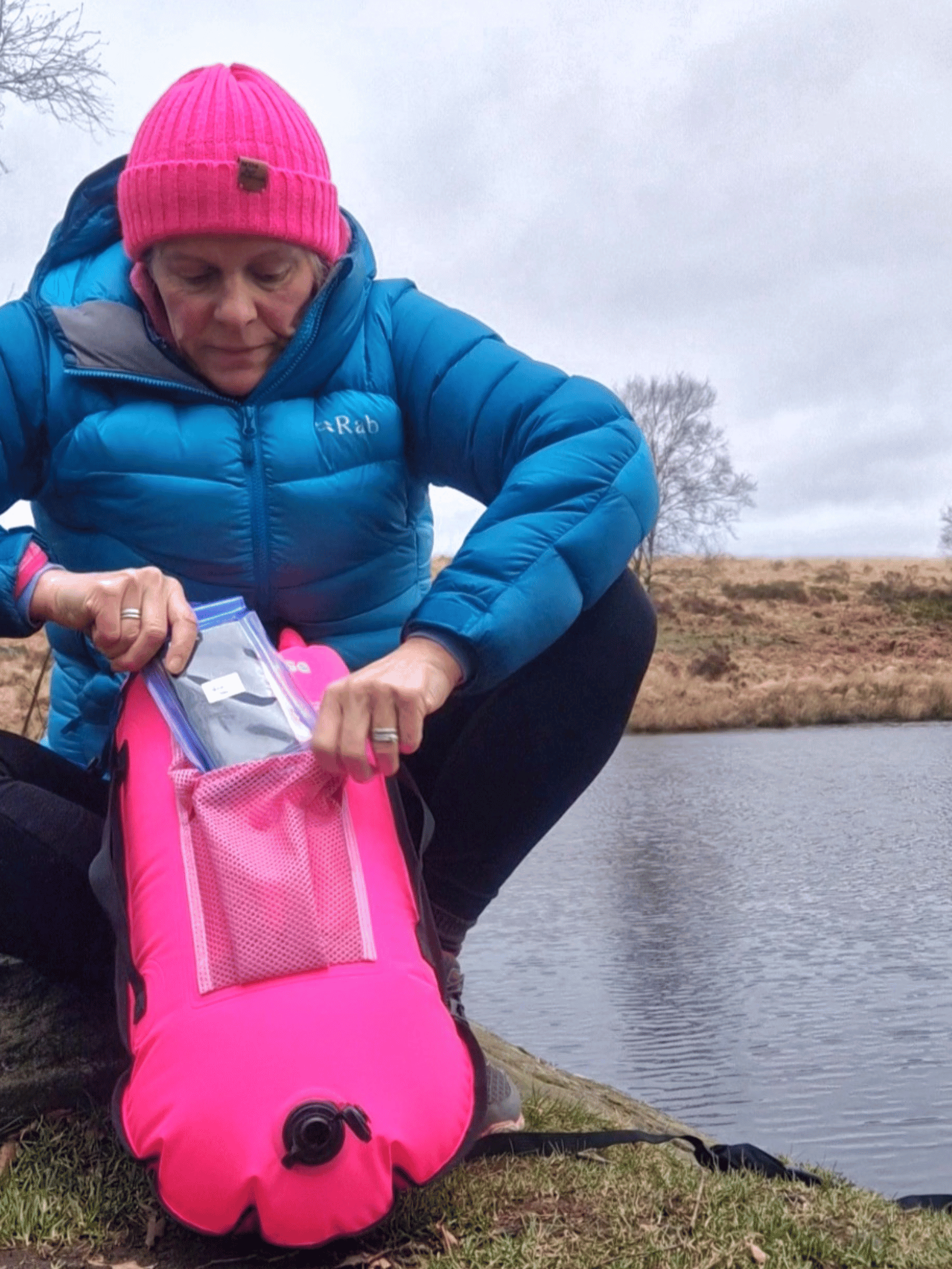 lady at the side of a lake putting a waterproof phone case into the mesh pocket on top of a pink tow float