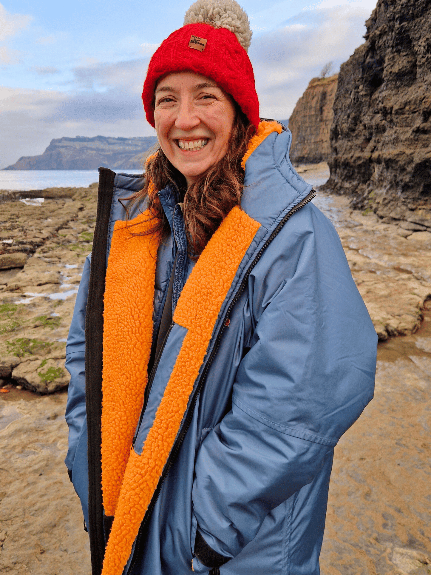 lady by the sea wearing a blue changing robe with bright orange lining. She is smiling broadly