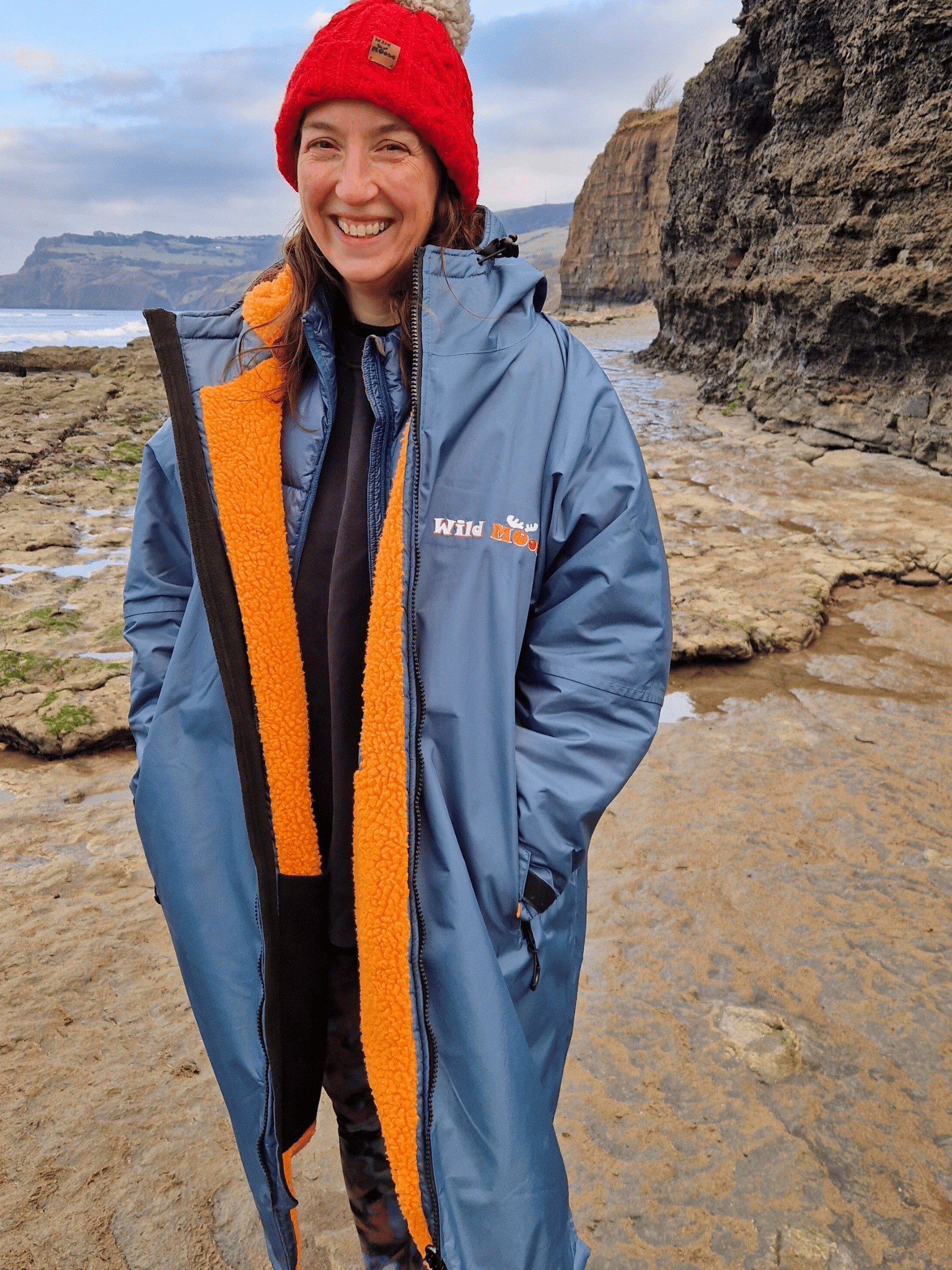 lady by the sea wearing a blue changing robe with bright orange lining. She is smiling broadly