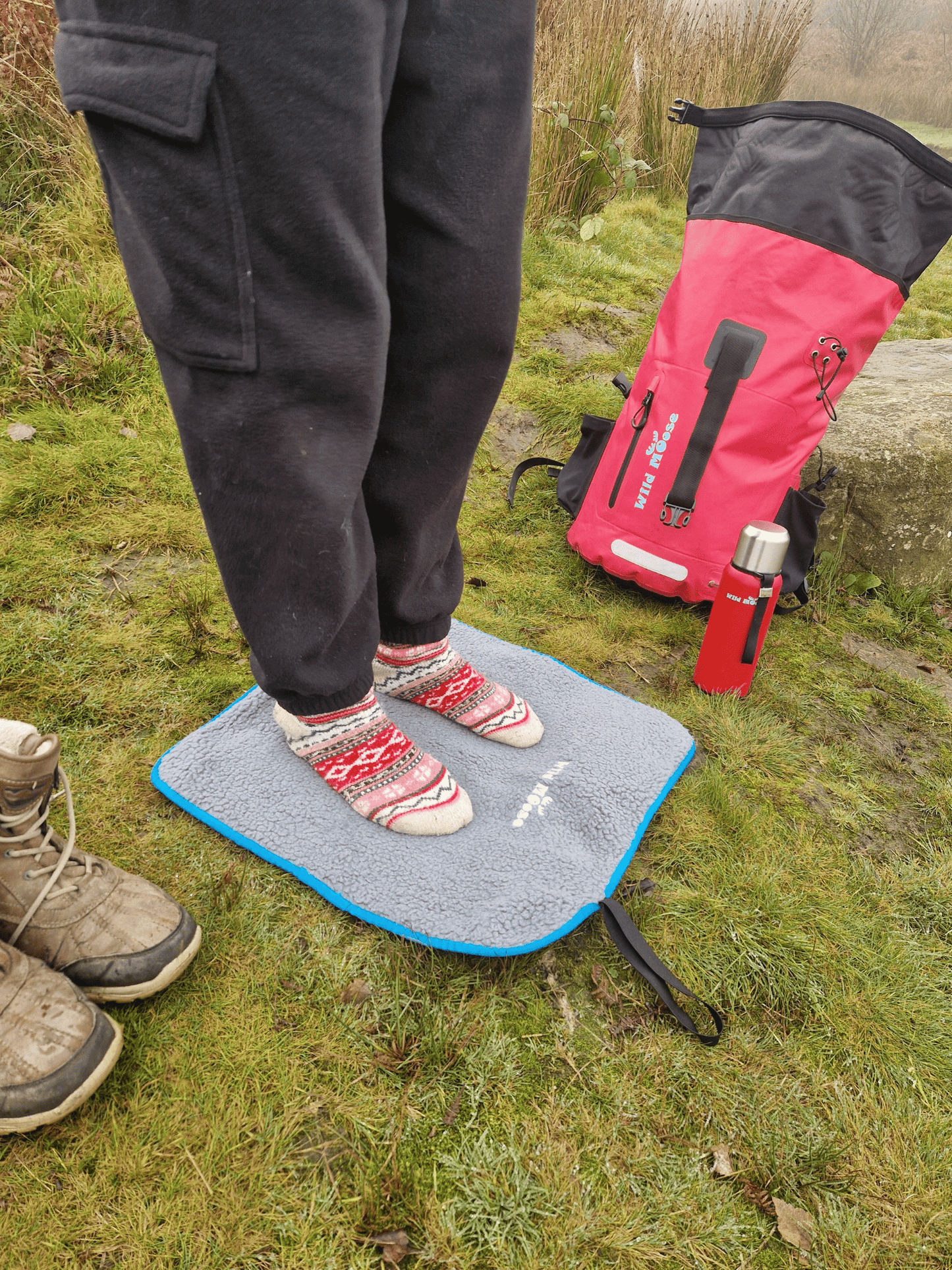 socked feet on a fleece lined changing mat on grass, with a pair of boots next to the mat and a red flask and pink backpack in the background