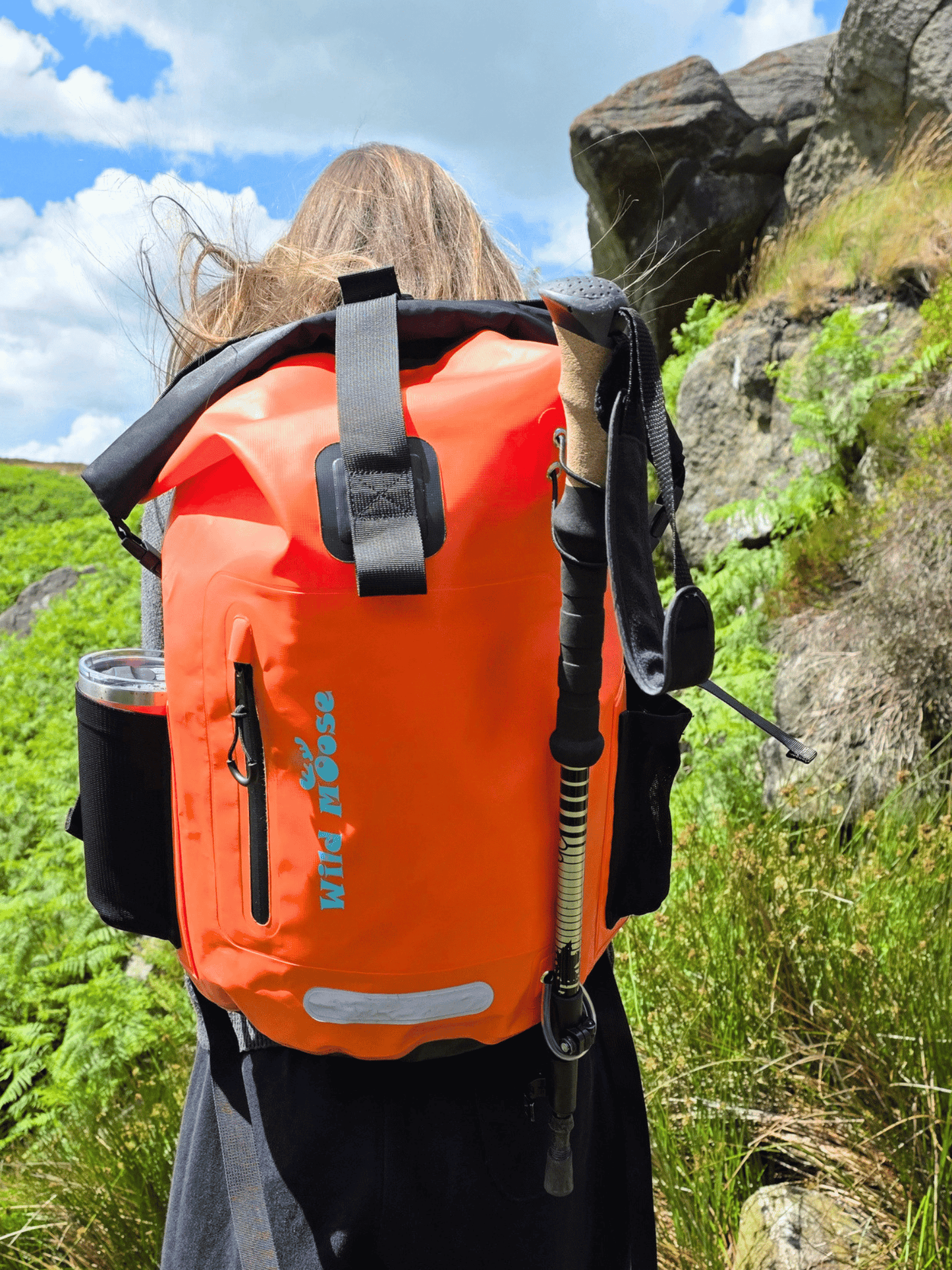 An outdoor shot of a person from behind wearing a bright orange waterproof backpack with black straps and gear, including a trekking pole and side cup holder, set against a natural rocky and green background.