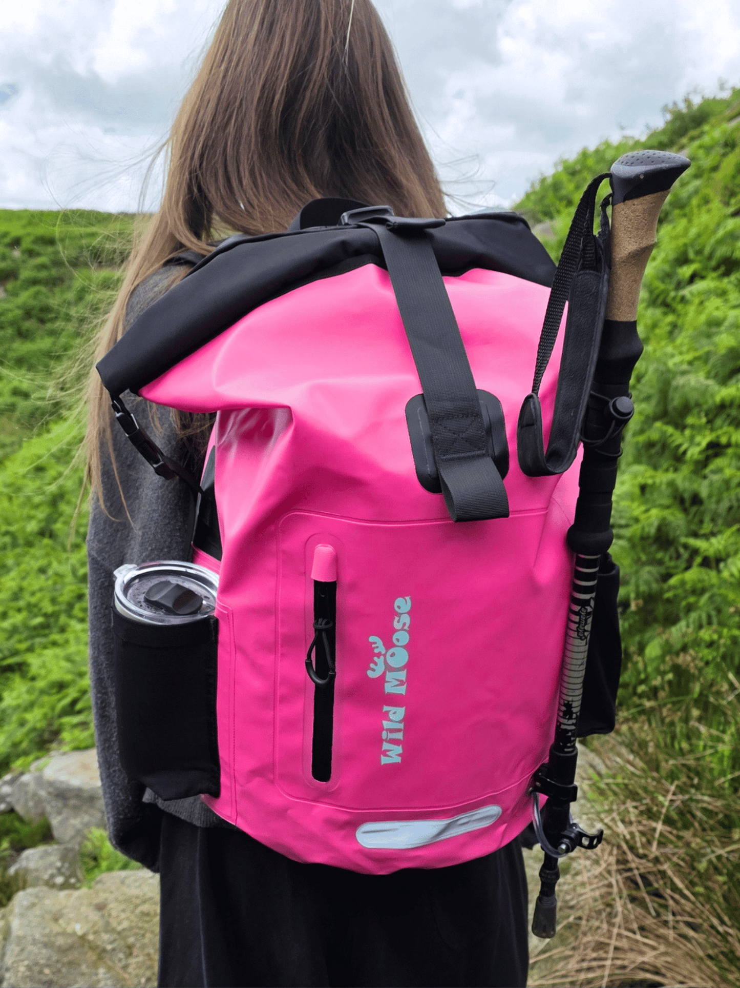 A bright pink waterproof roll-top backpack with black straps and a black vertical zippered pocket, shown from the back on a person outdoors. The backpack has 'WILD MOOSE' printed vertically in teal on the front pocket, and a black hiking pole is tucked into the side pocket.