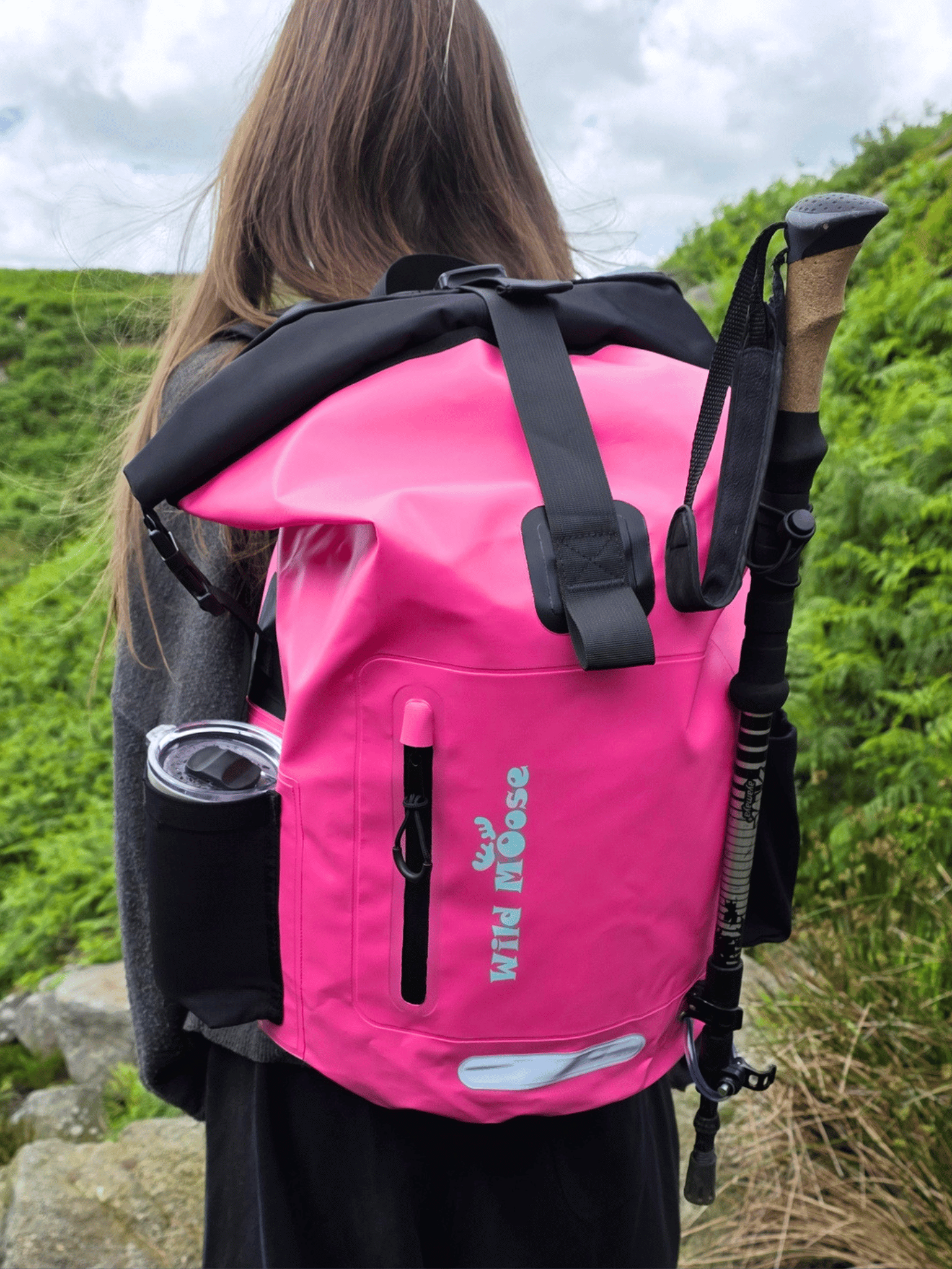 A bright pink waterproof roll-top backpack with black straps and a black vertical zippered pocket, shown from the back on a person outdoors. The backpack has 'WILD MOOSE' printed vertically in teal on the front pocket, and a black hiking pole is tucked into the side pocket.