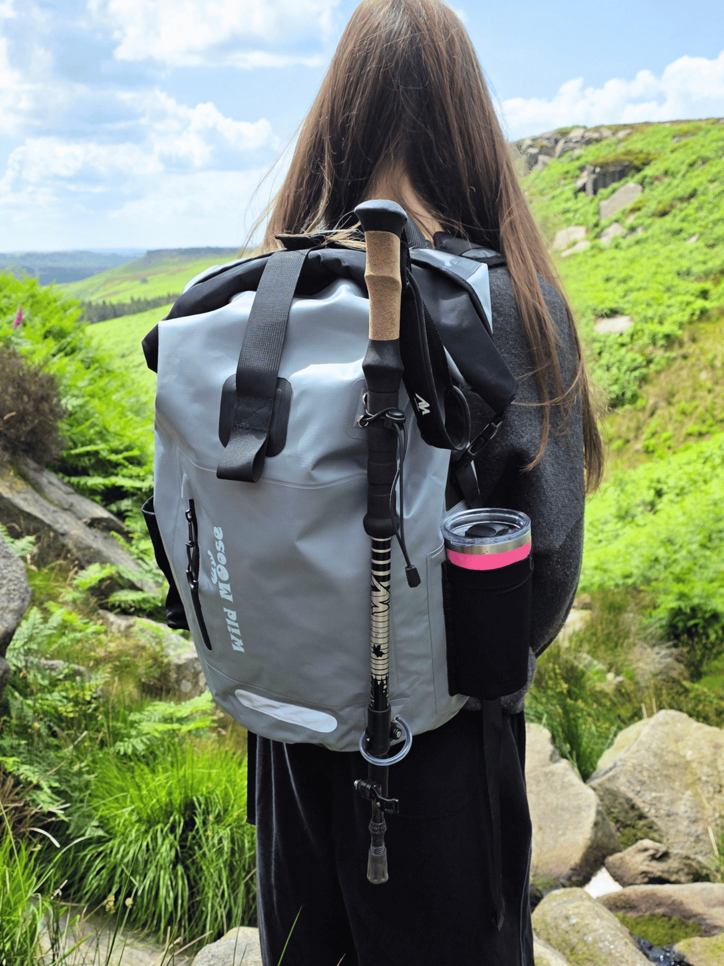 A person viewed from behind wearing a gray waterproof roll-top backpack with black straps and a black side cup holder containing a pink-tinted bottle, standing outdoors on a rocky green hillside.