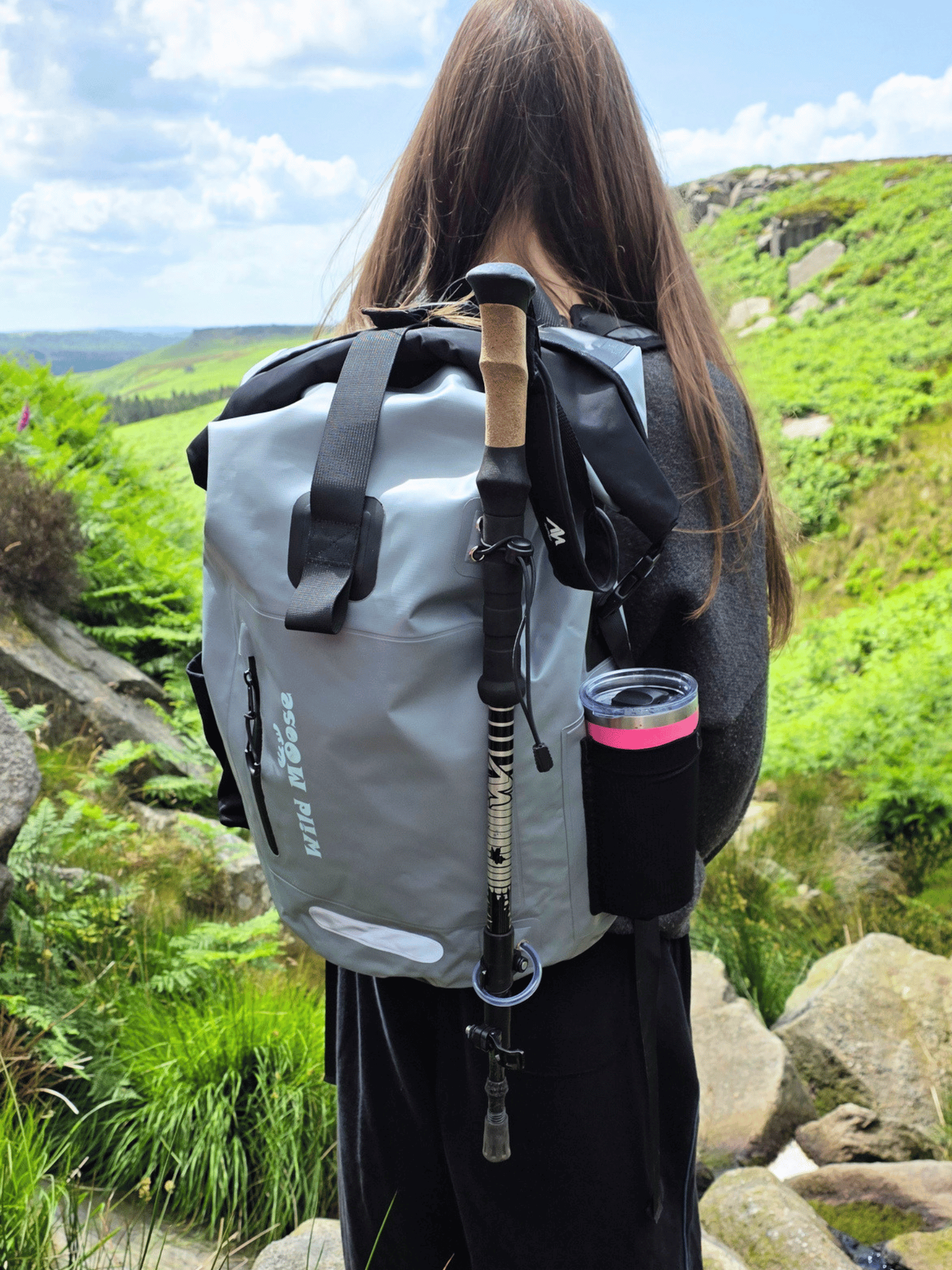 A person viewed from behind wearing a gray waterproof roll-top backpack with black straps and a black side cup holder containing a pink-tinted bottle, standing outdoors on a rocky green hillside.