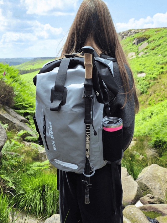 A person viewed from behind wearing a gray waterproof roll-top backpack with black straps and a black side cup holder containing a pink-tinted bottle, standing outdoors on a rocky green hillside.