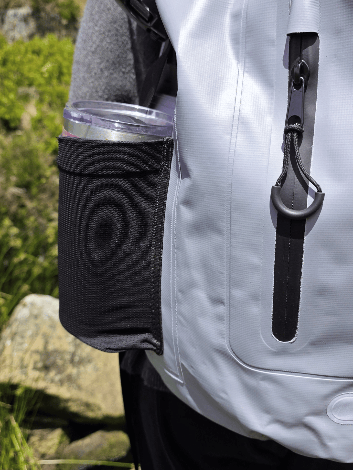 close up picture of a black splashproof zip pocket and cup holder worn by a person stood in a rocky grassy landscape.