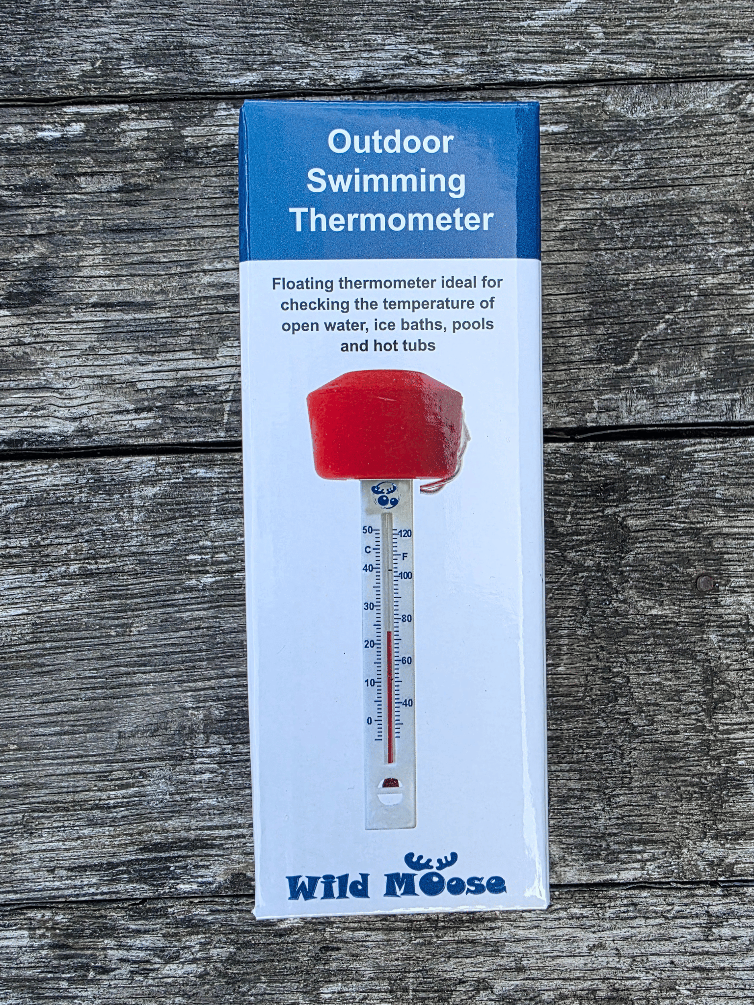 Packaged outdoor swimming thermometer by Wild Moose, featuring an orange float with a white thermometer tube printed on the box, set against a weathered grey wood background.