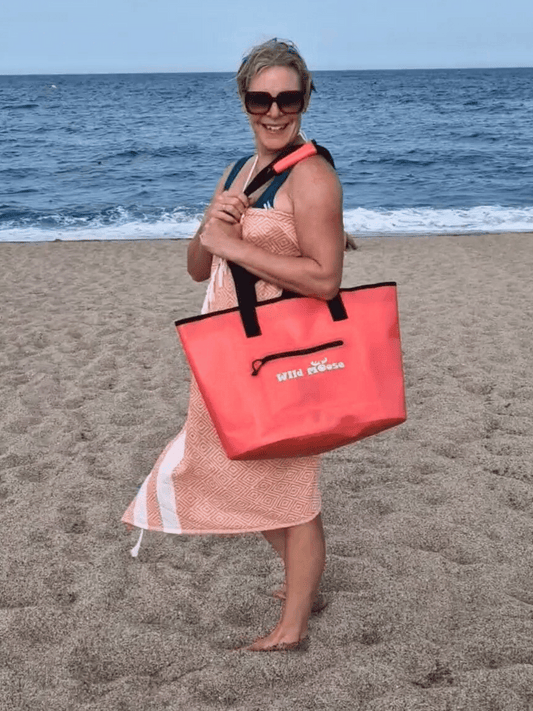 lady smiling on a beach in a hammam towel, with a coral-coloured Wild Moose tote bag over her shoulder