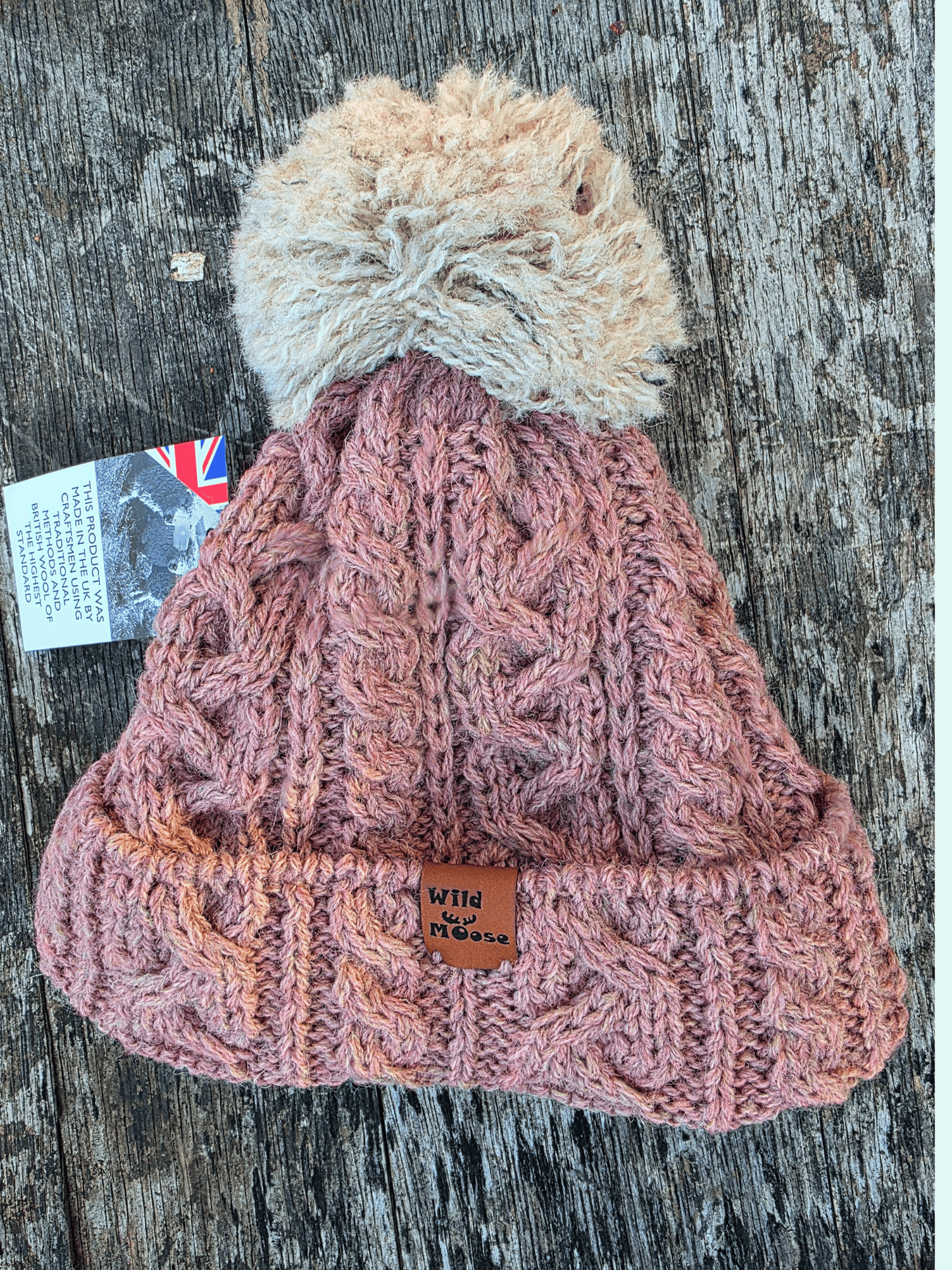 Dusty pink knitted bobble hat with a beige pom-pom on a wooden surface