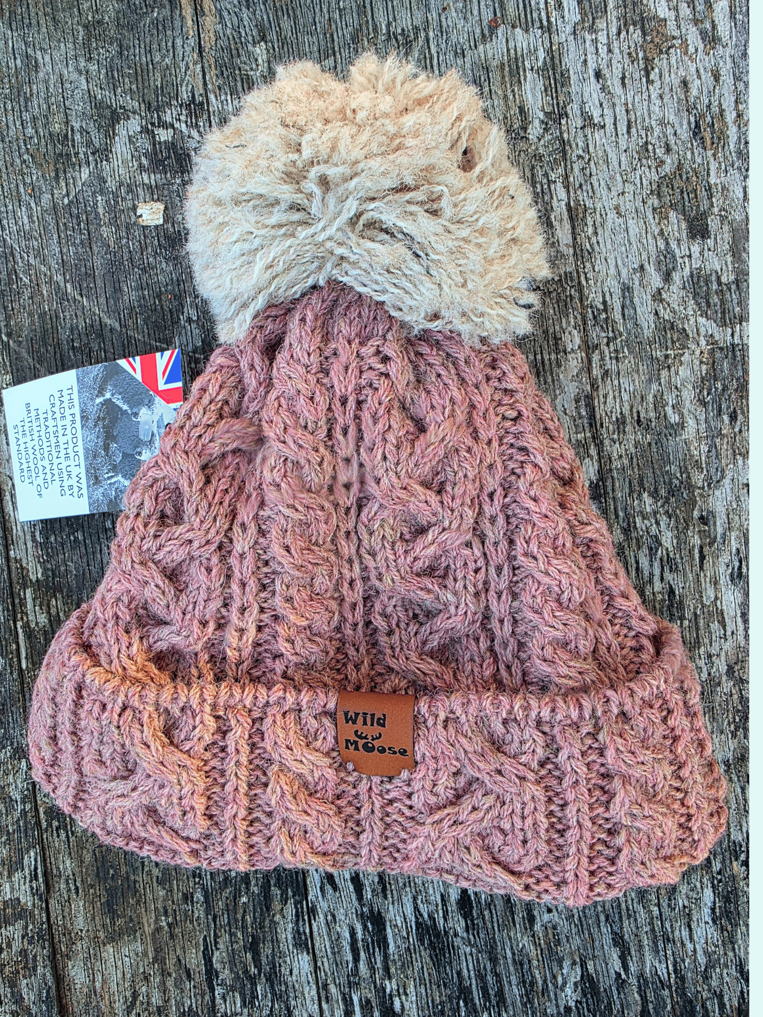 Dusty pink knitted bobble hat with a beige pom-pom on a wooden surface