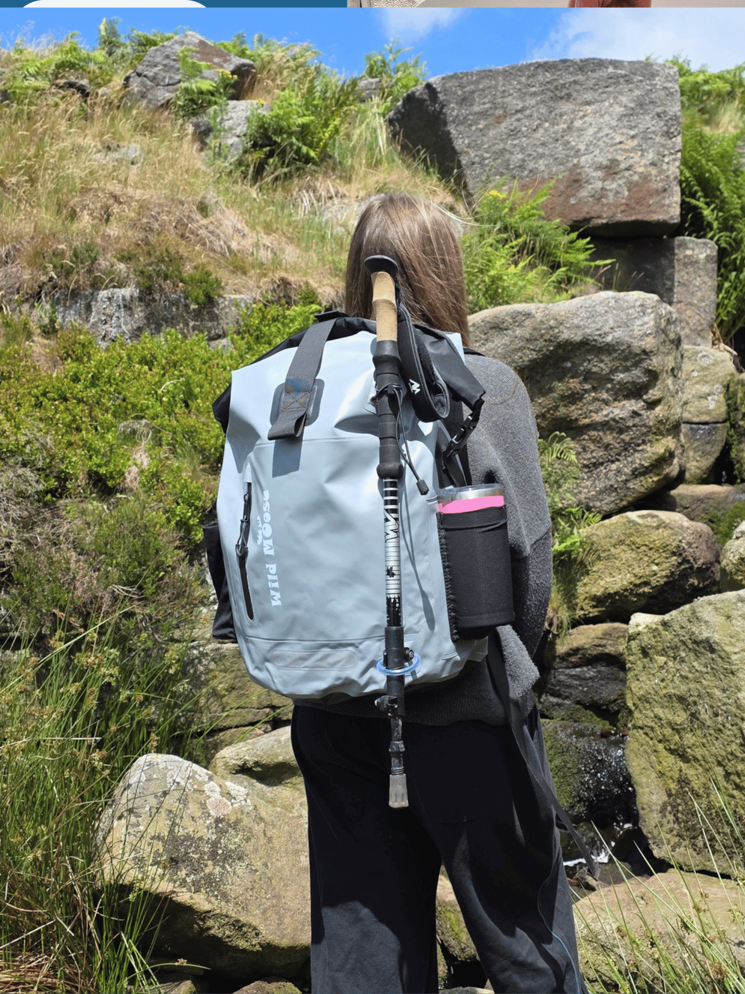 A person outdoors on a rocky hillside wearing a light gray waterproof backpack with a black hiking pole tucked into the black hiking pole loops and a black side pouch containing a pink flask.
