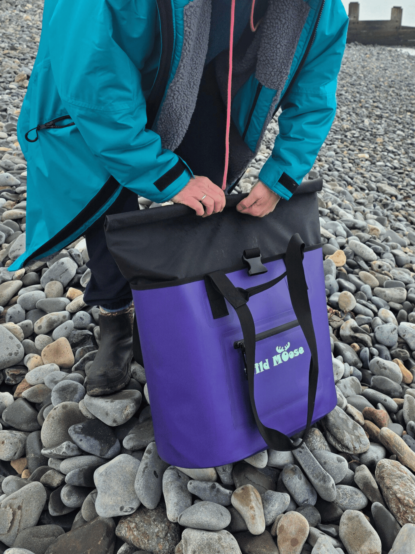 Person unfolding the black top of a purple tote bag on a pebbly beach