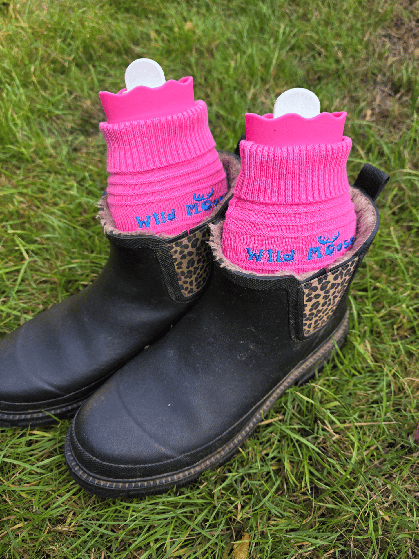 2 bright pink mini hot water bottles in a knitted cover in a pair of ankle length black wellies on grassy ground