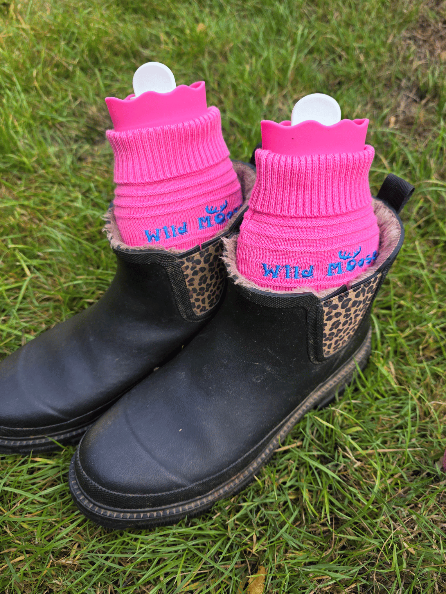 2 bright pink mini hot water bottles in a knitted cover in a pair of ankle length black wellies on grassy ground