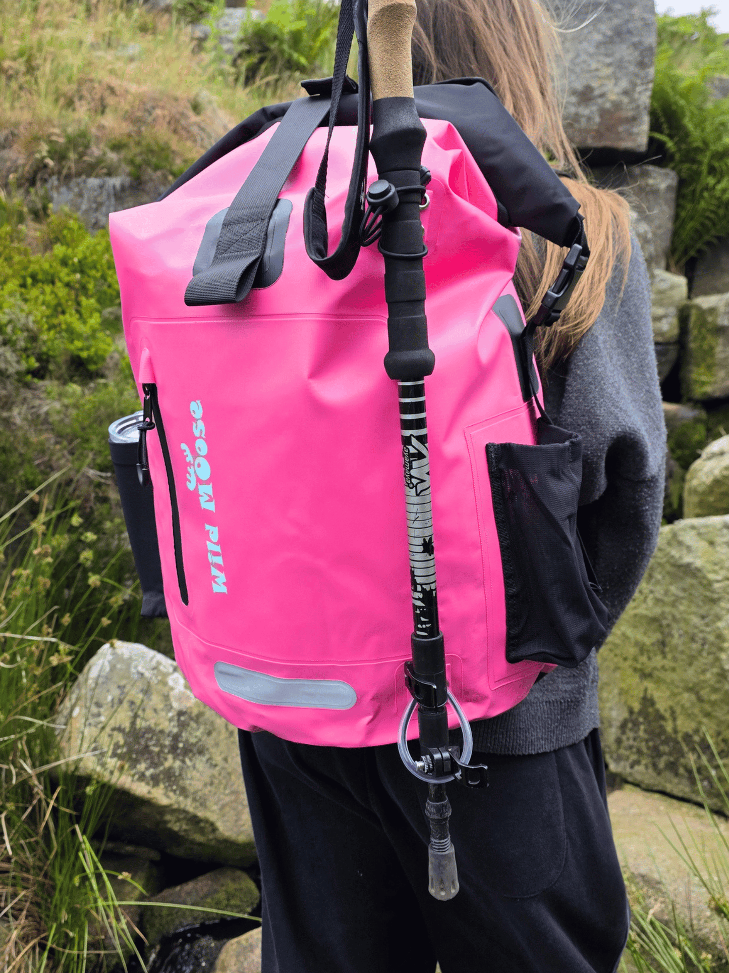 A bright pink waterproof backpack with black straps and a vertical black-and-white patterned walking pole, shown worn by a person outdoors among rocks and grass. The backpack has visible 'Wild Moose' branding.