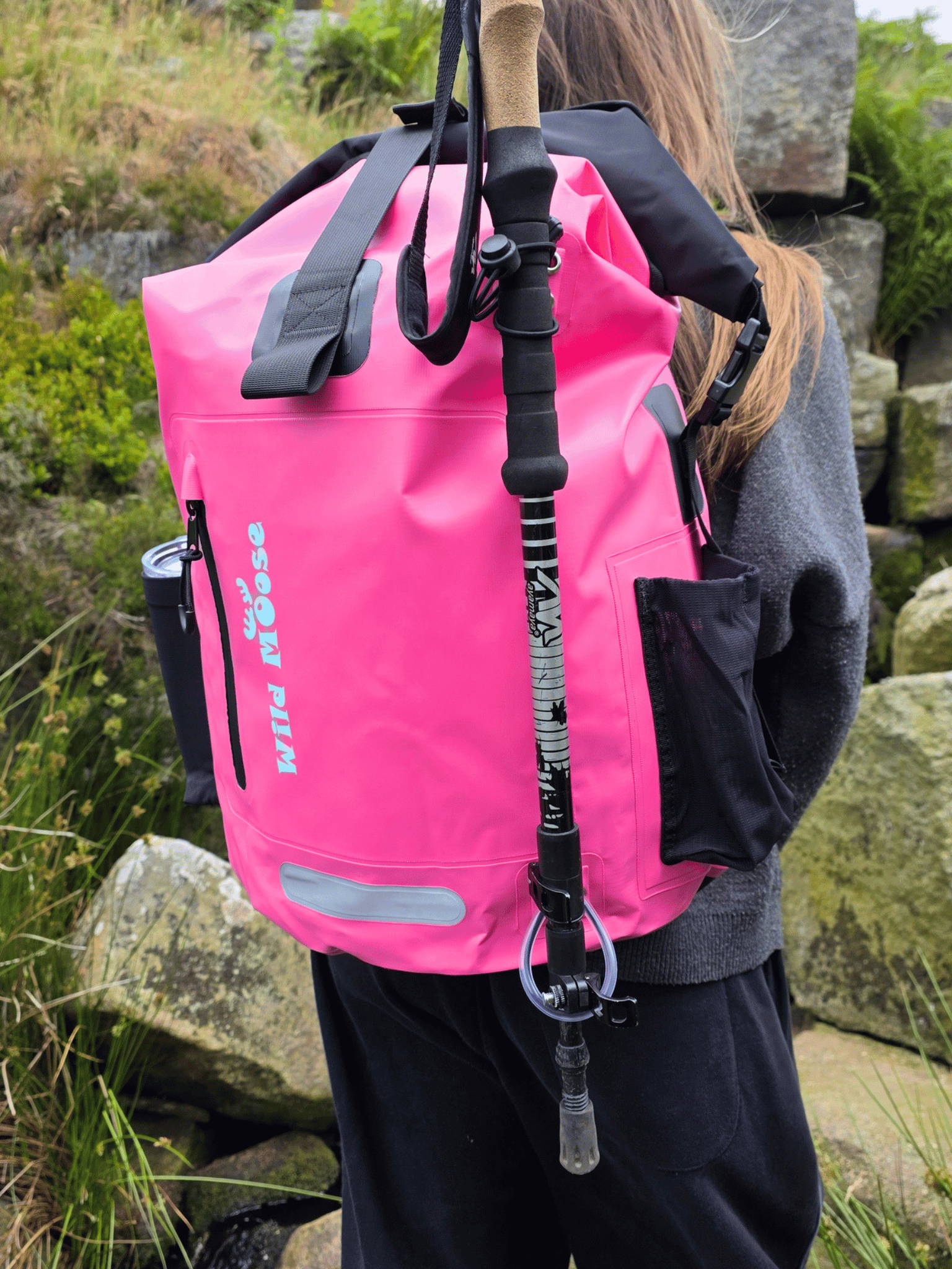 A bright pink waterproof backpack with black straps and a vertical black-and-white patterned walking pole, shown worn by a person outdoors among rocks and grass. The backpack has visible 'Wild Moose' branding.