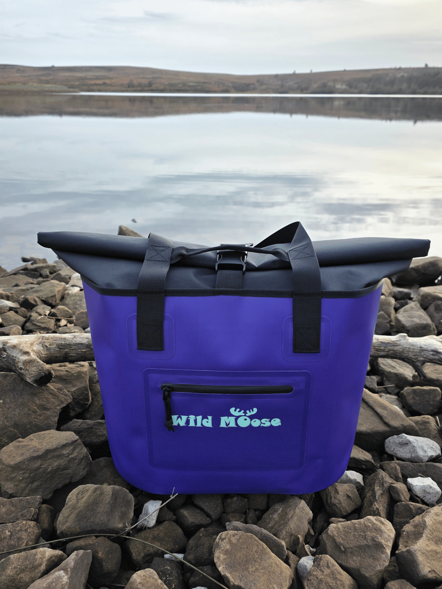 Purple tote bag with black straps on a rocky shore with water in the background
