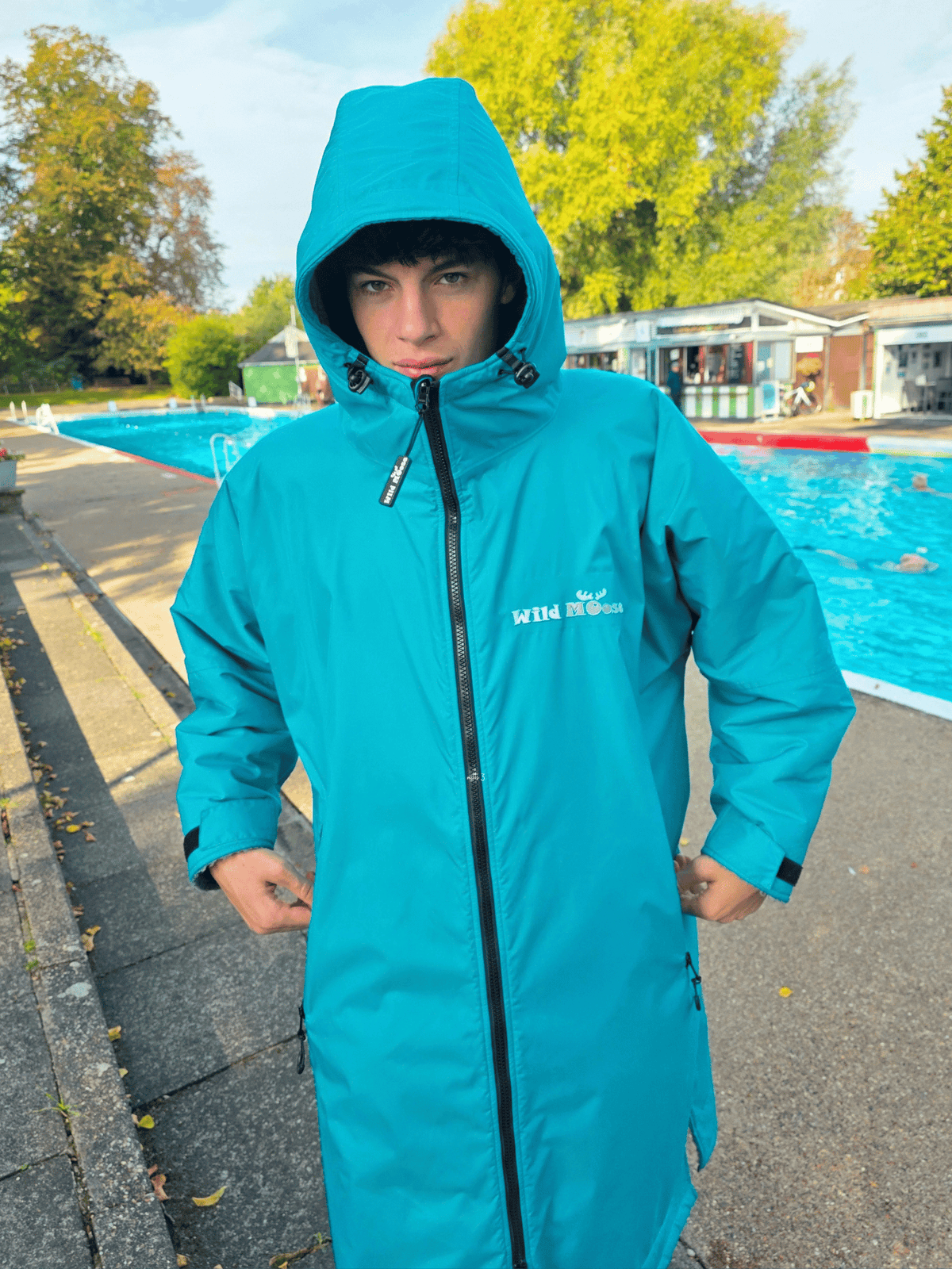 A person wearing a bright teal hooded changing robe with a black front zipper, standing outdoors beside an outdoor swimming pool. The coat has a white printed logo on the left chest reading 'Wild Moose'.