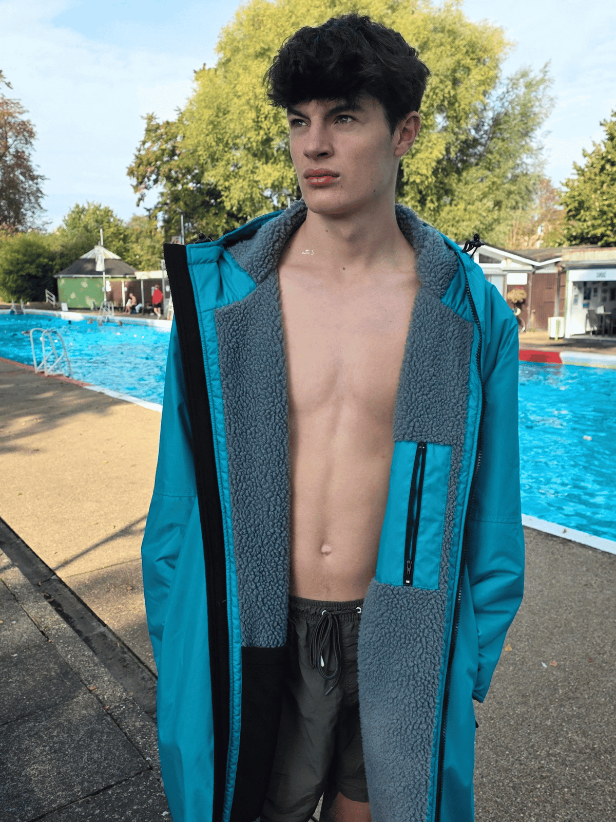 A young man standing outdoors by a swimming pool wearing a bright teal and grey fleece-lined hoodedchanging robe open over his bare torso.