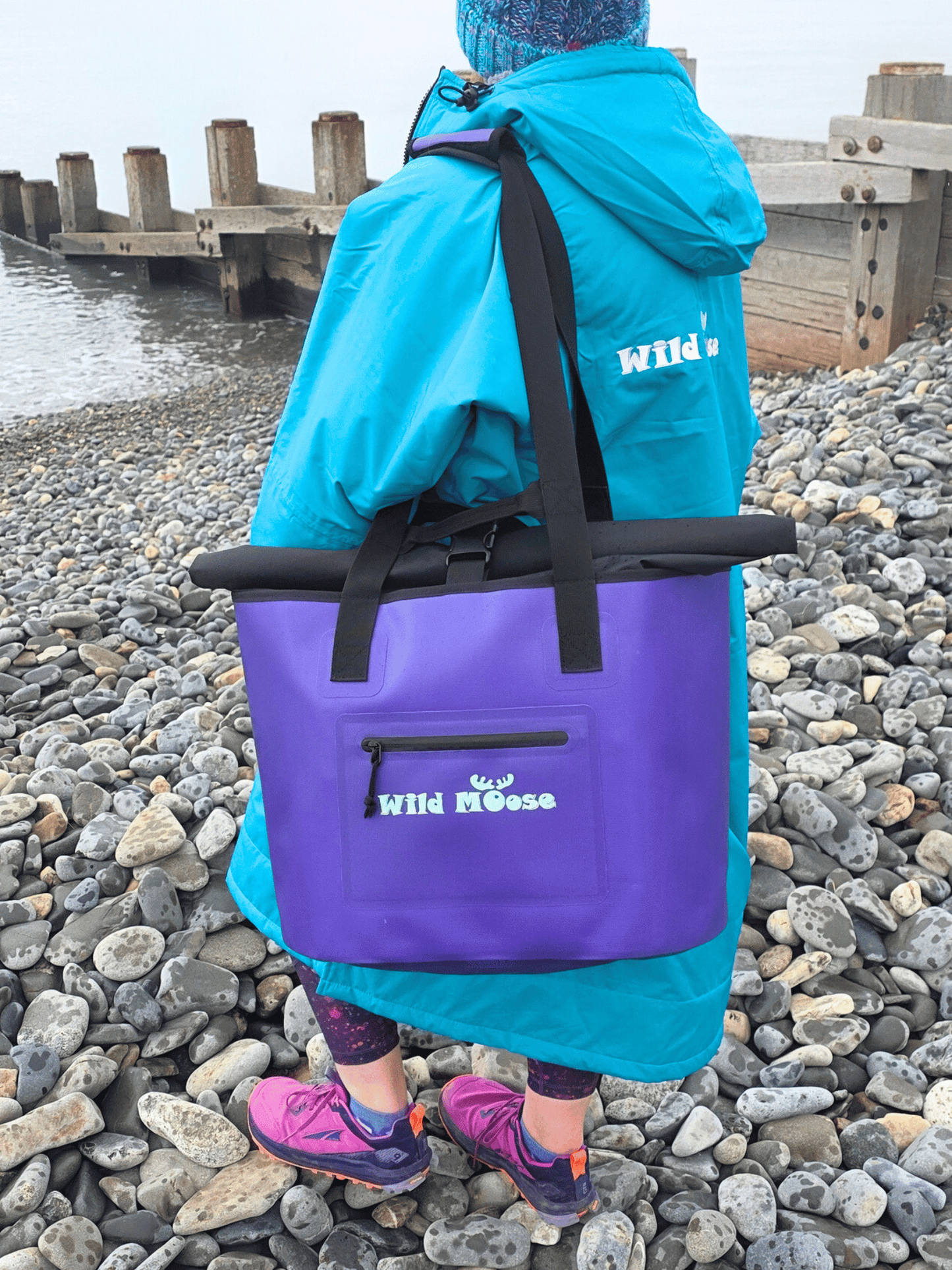 Person wearing a green/blue changing robe and carrying a purple tote bag with 'Wild Moose' branding on a pebbly beach with groynes in the background.