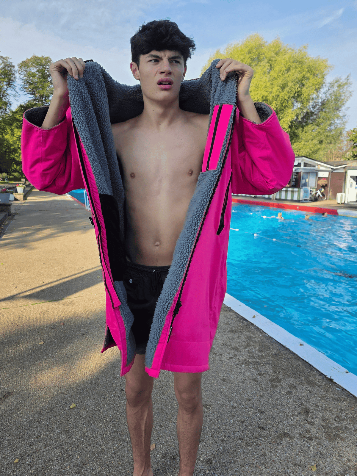 A shirtless young man standing outdoors by a swimming pool, holding open a bright pink fleece-lined jacket or robe with dark gray sherpa lining. The pool, trees, and buildings are visible in the background.