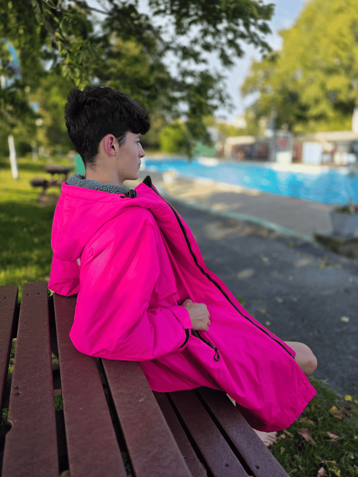 A man sitting on a wooden bench outdoors, wearing a bright hot pink hooded changing robe. The background shows a blurred outdoor setting with trees, a pool, and a pathway.
