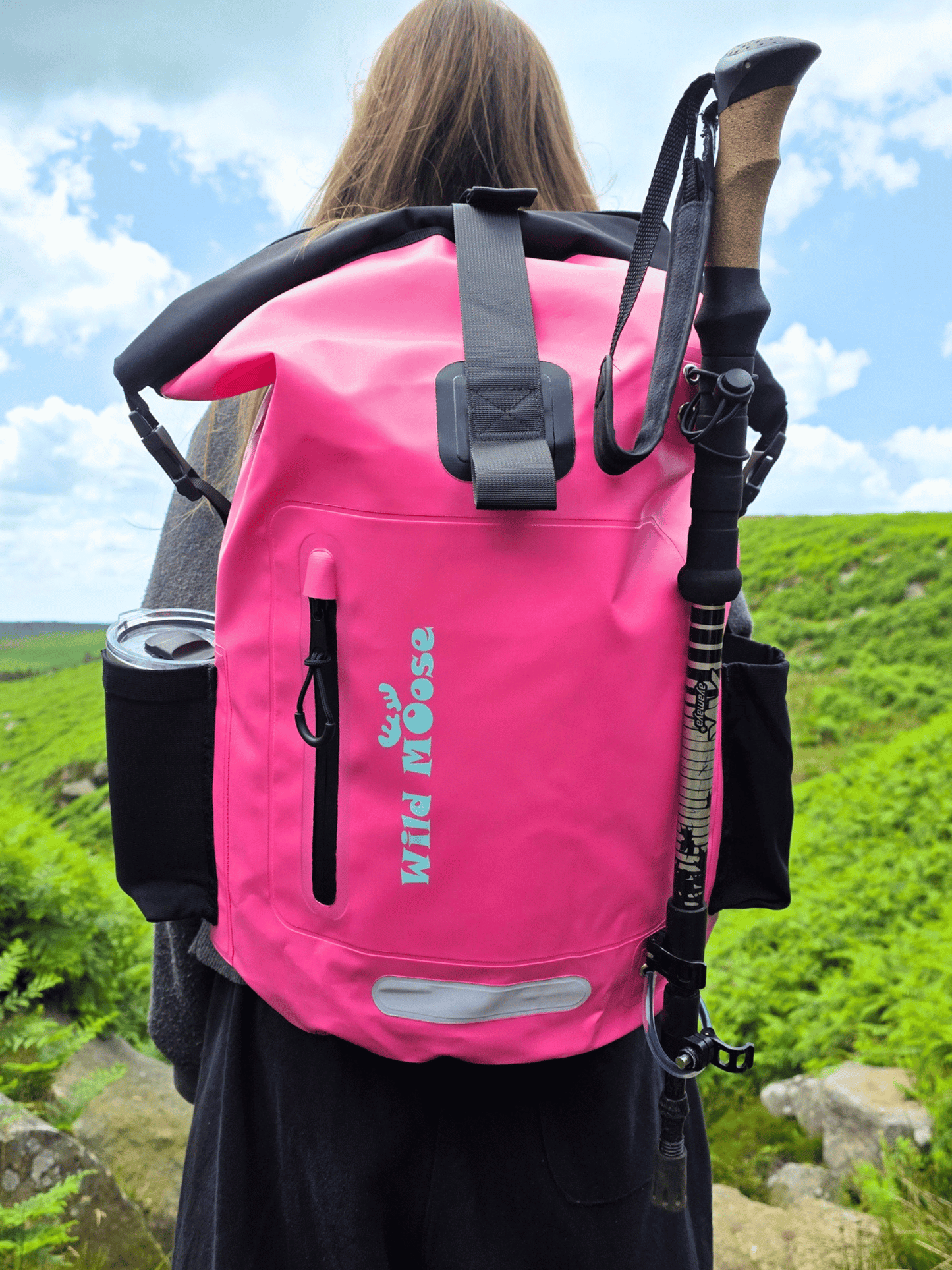 A bright pink waterproof backpack with black straps and gear attachments, shown from behind on a person outdoors in a grassy landscape. The backpack has the brand name 'Wild Moose' printed vertically on the front.