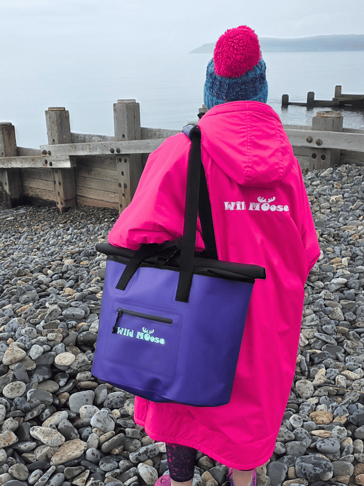Person wearing a pink robe and holding a purple tote bag with 'Wild Moose' branding on a pebbly beach.