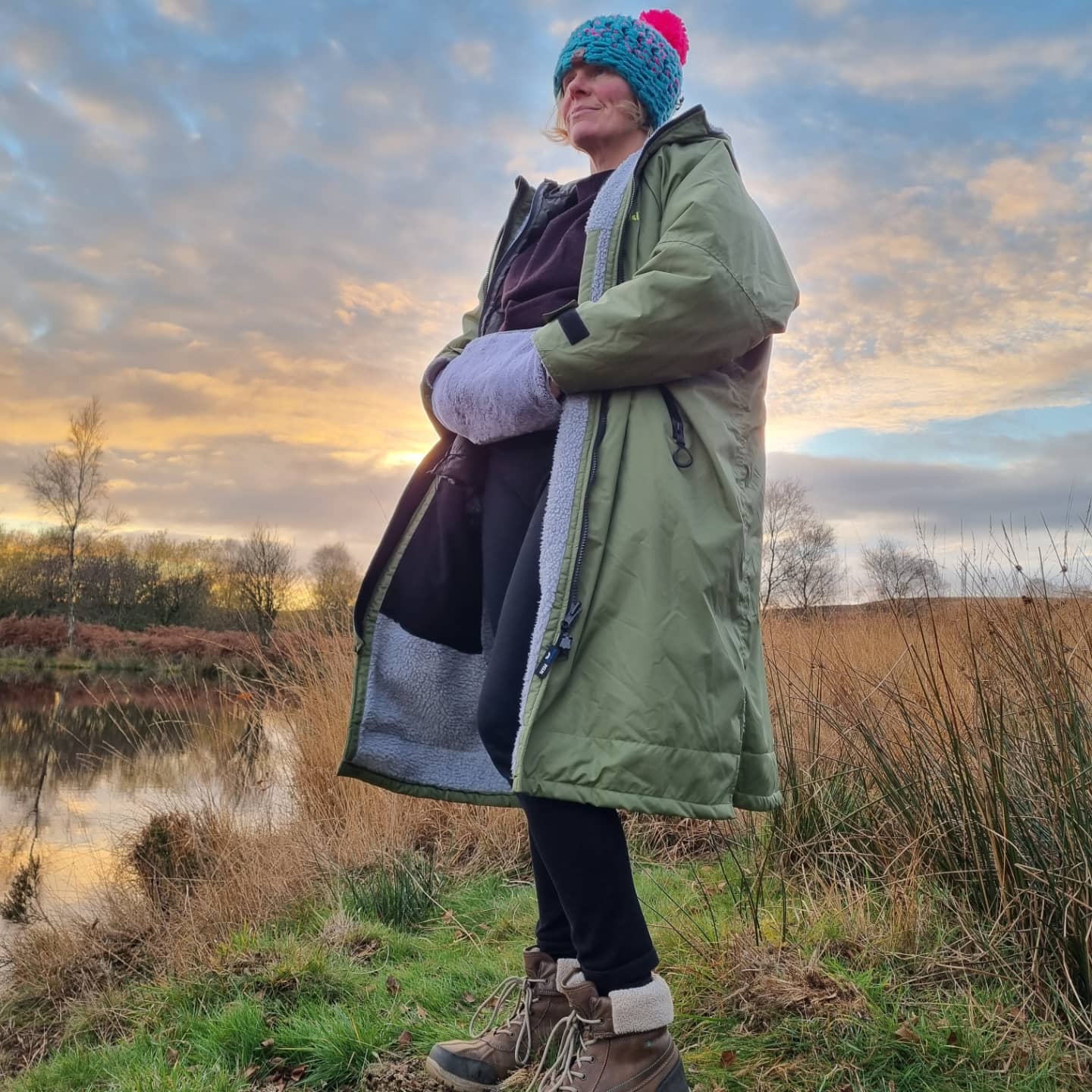 a lady is standing by a lake and the sun is setting in the sky. She is wearing the Hottum core warmer and has her hands warming in it. She is also wearing a khaki Wild Moose changing robe and  a turquoise and pink Big Moose Bobble hat