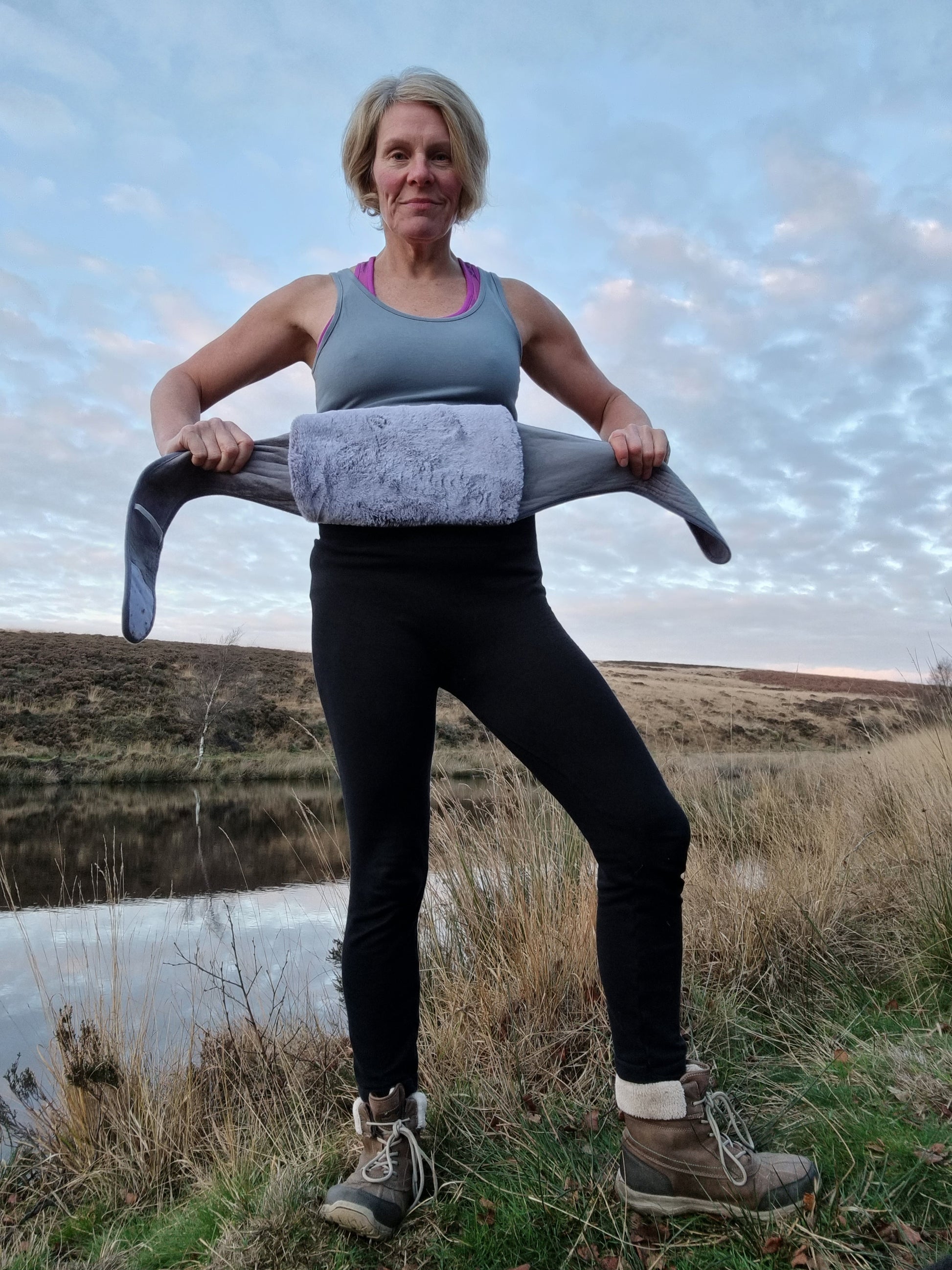 a lady is standing in front of a lake and holding a grey Hottum, a core warmer made of faux fur. She is holding it be the straps to show how it attaches simply behind your back
